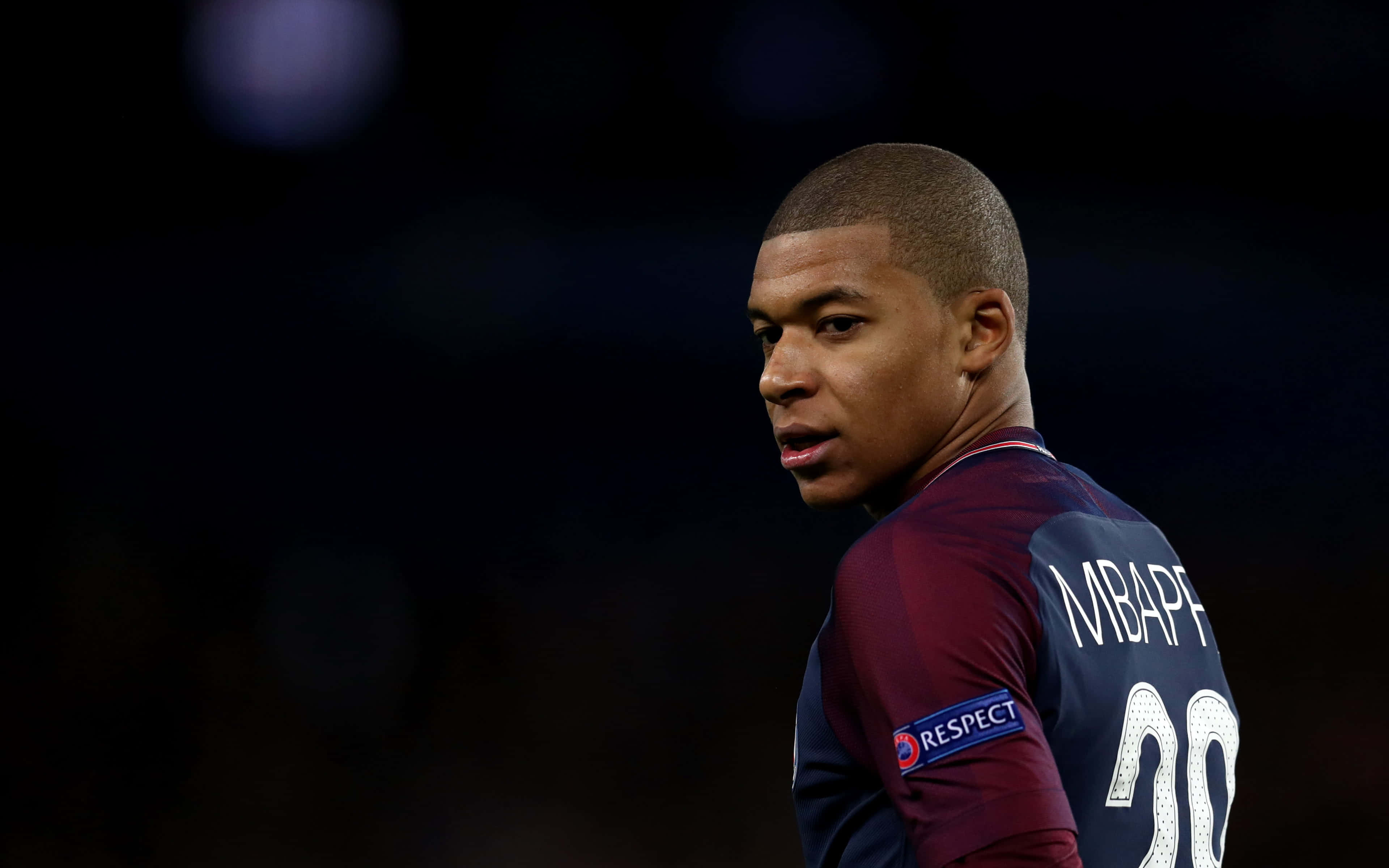 Mbappe 桌布 壁紙 — 身穿栗色球衣的足球員基利安·麥巴比在黑暗模糊的球場背景下回頭看的特寫。