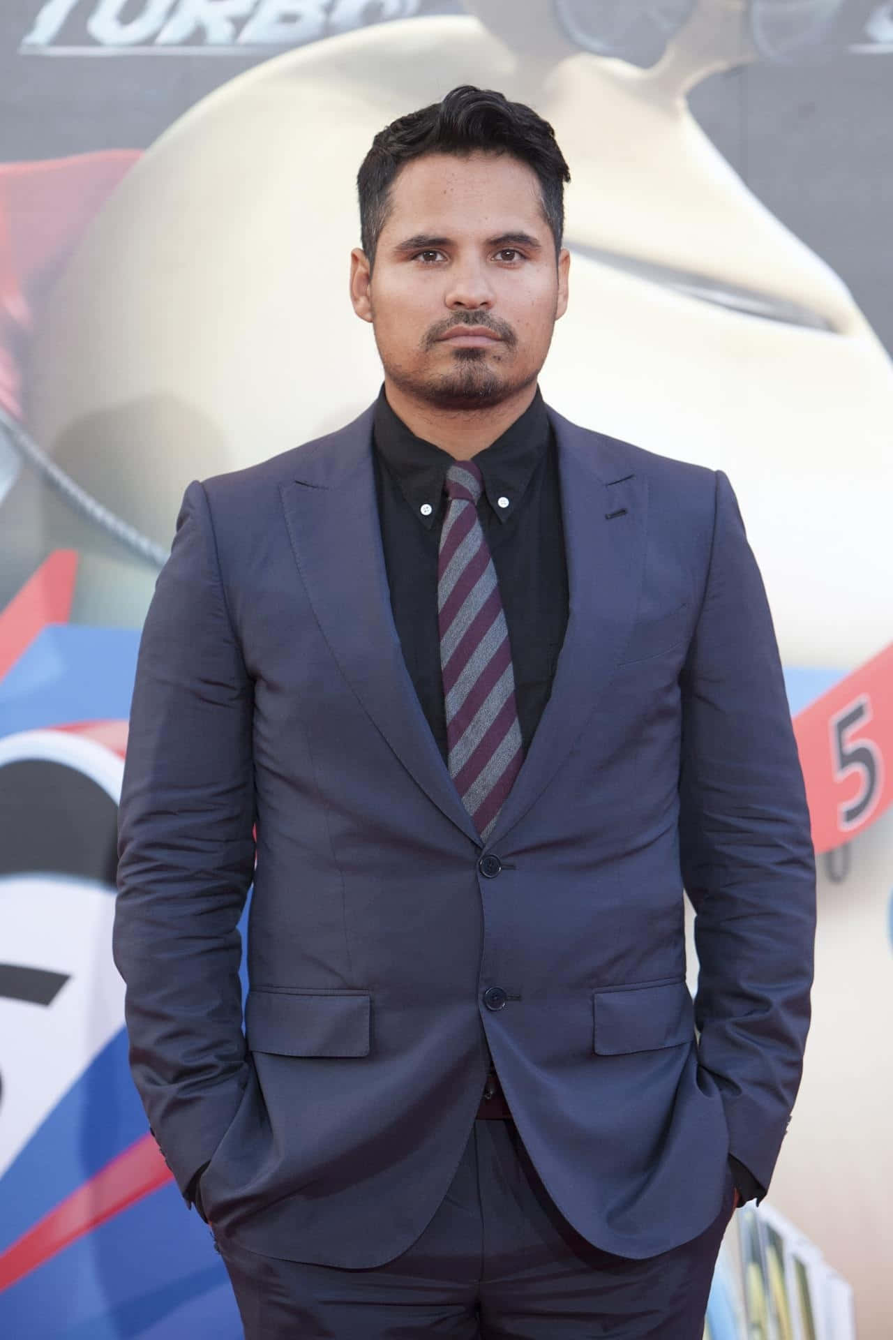 Michael Peña 桌布 壁紙 — 身穿海軍藍西裝和條紋領帶的男子自信地站在電影首映禮上，雙手插在口袋裡。