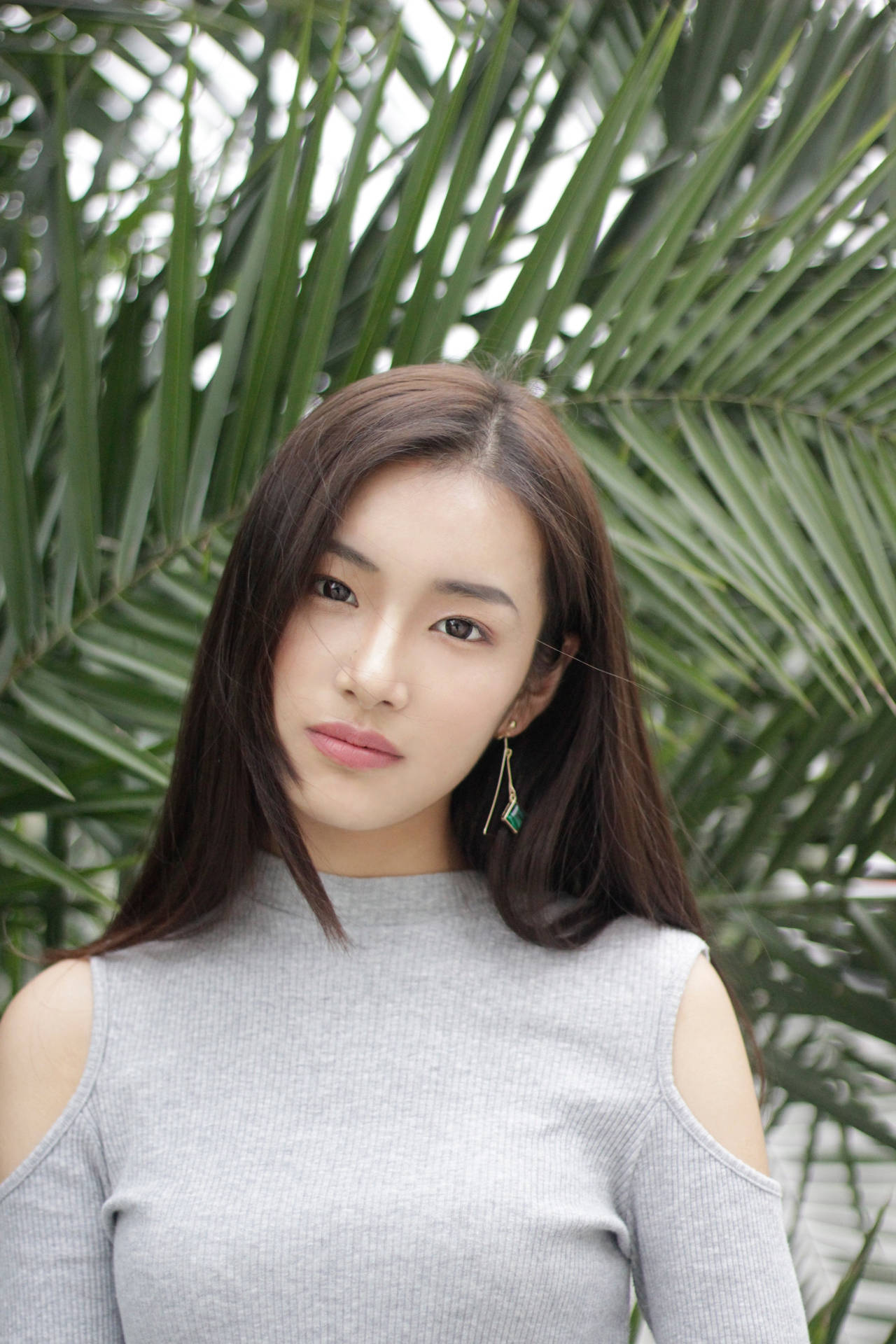 身穿灰色上衣、戴著幾何耳環的年輕女性站在棕櫚葉之間。