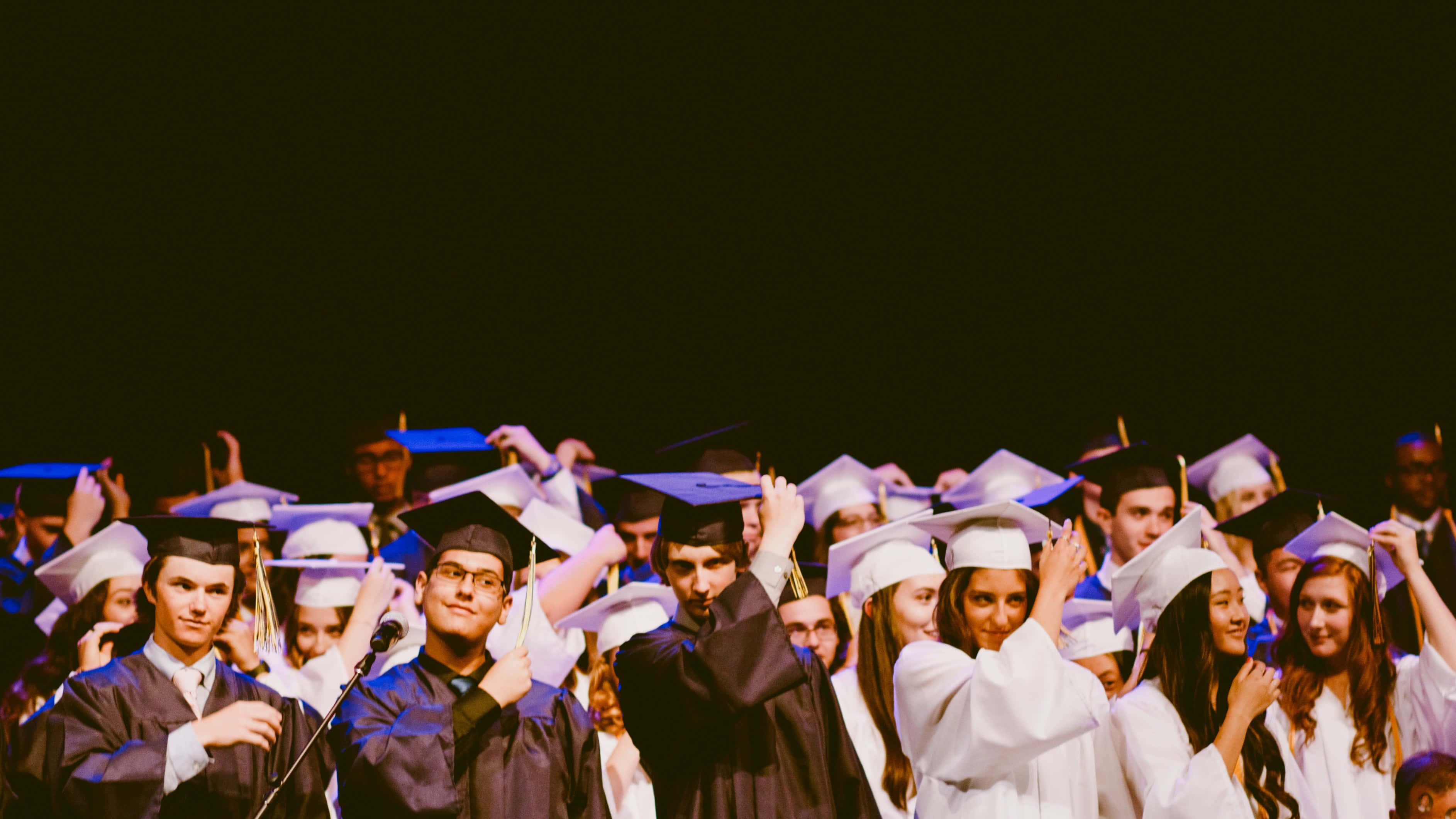 Graduation Ceremony 桌布 壁紙 — 身穿畢業袍和四方帽的畢業生在昏暗燈光下，於舞台上高舉雙手，帶著燦爛笑容歡樂慶祝。