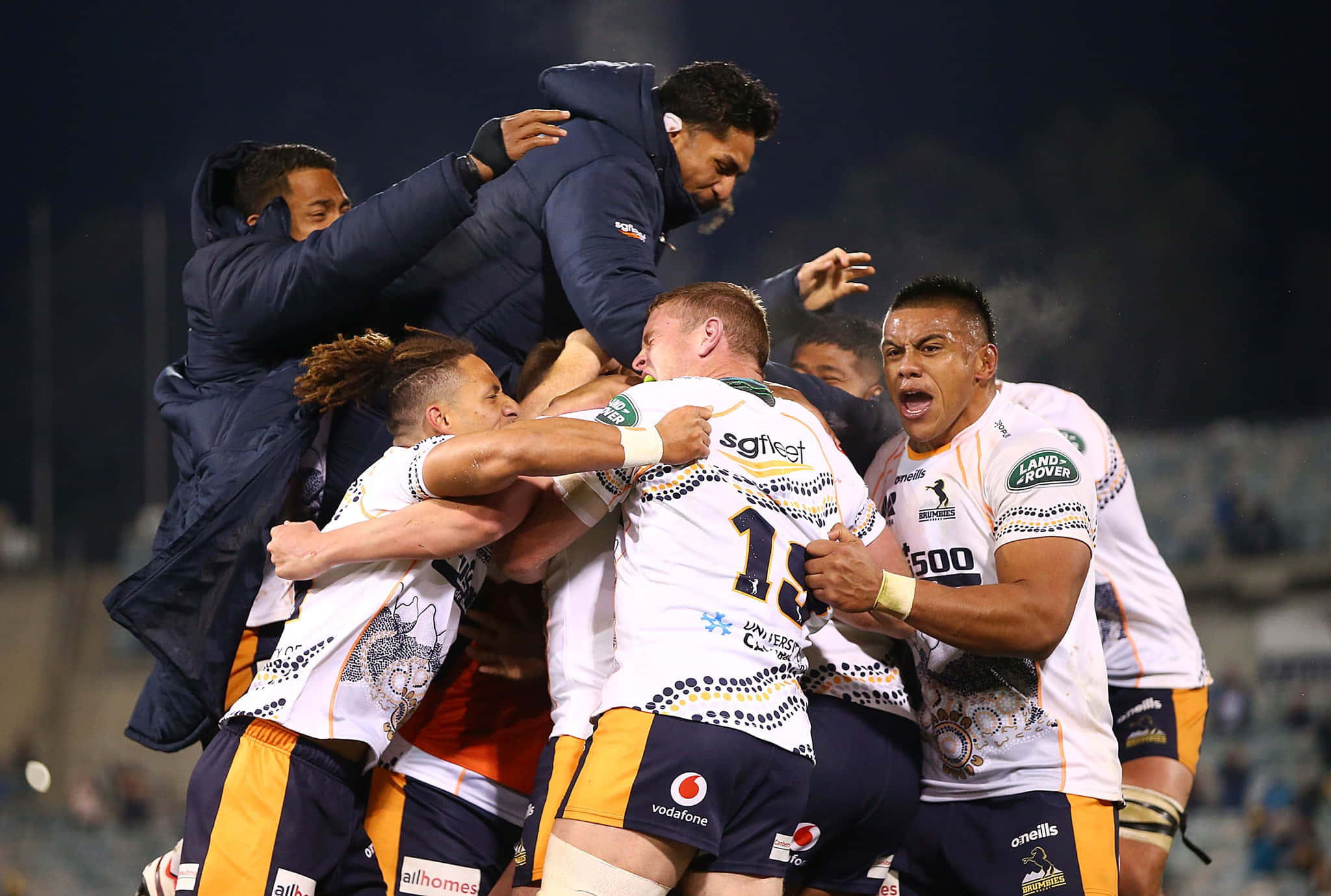 Brumbies 桌布 壁紙 — 身穿白色和橙色球衣的欖球運動員在夜間球場燈光下緊密圍成一圈慶祝勝利。