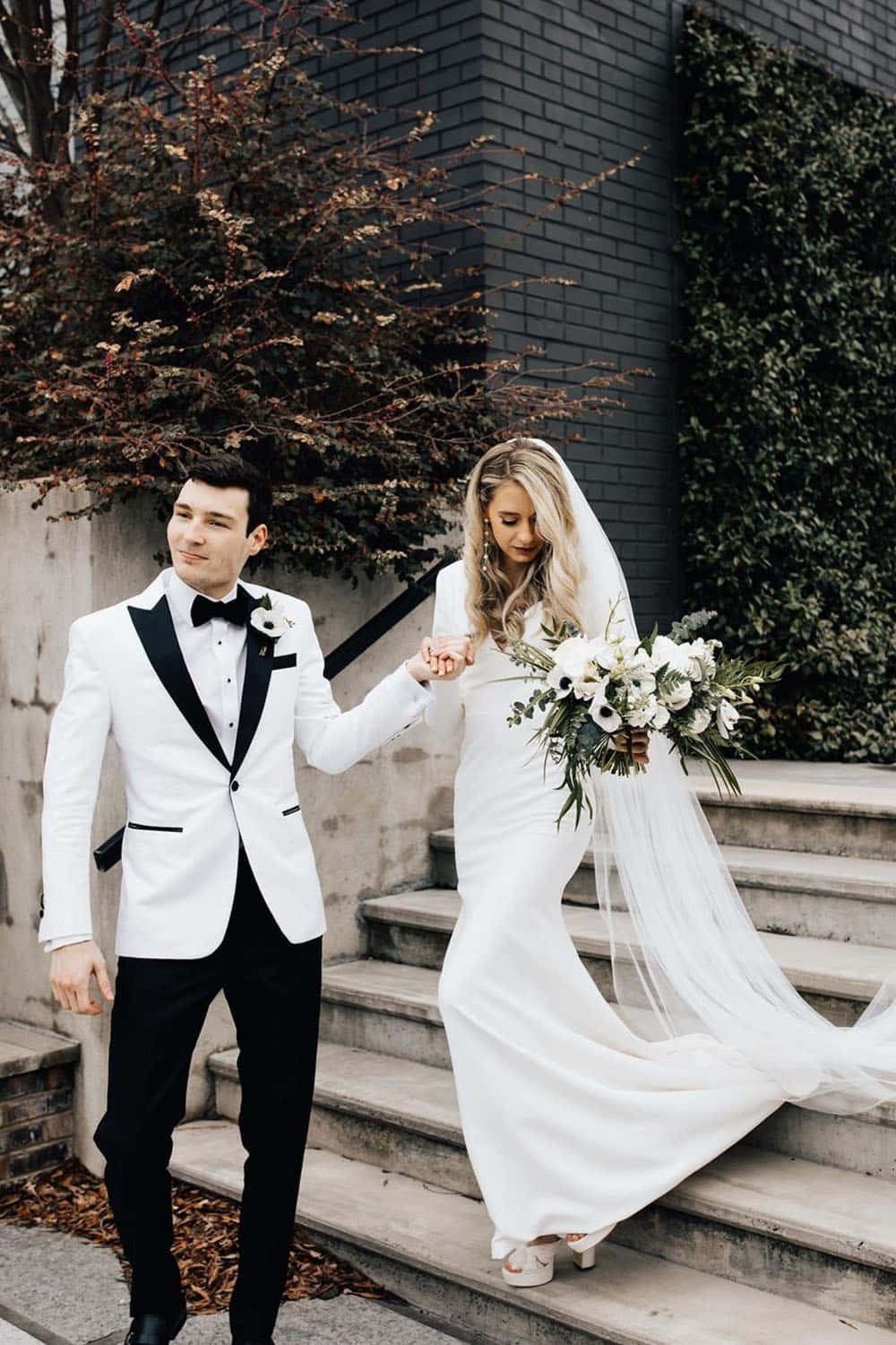 Black And White Wedding 桌布 壁紙 — 身穿白色和黑色正式服裝的新娘和新郎，沿著深色磚牆和秋葉旁的石階走下。