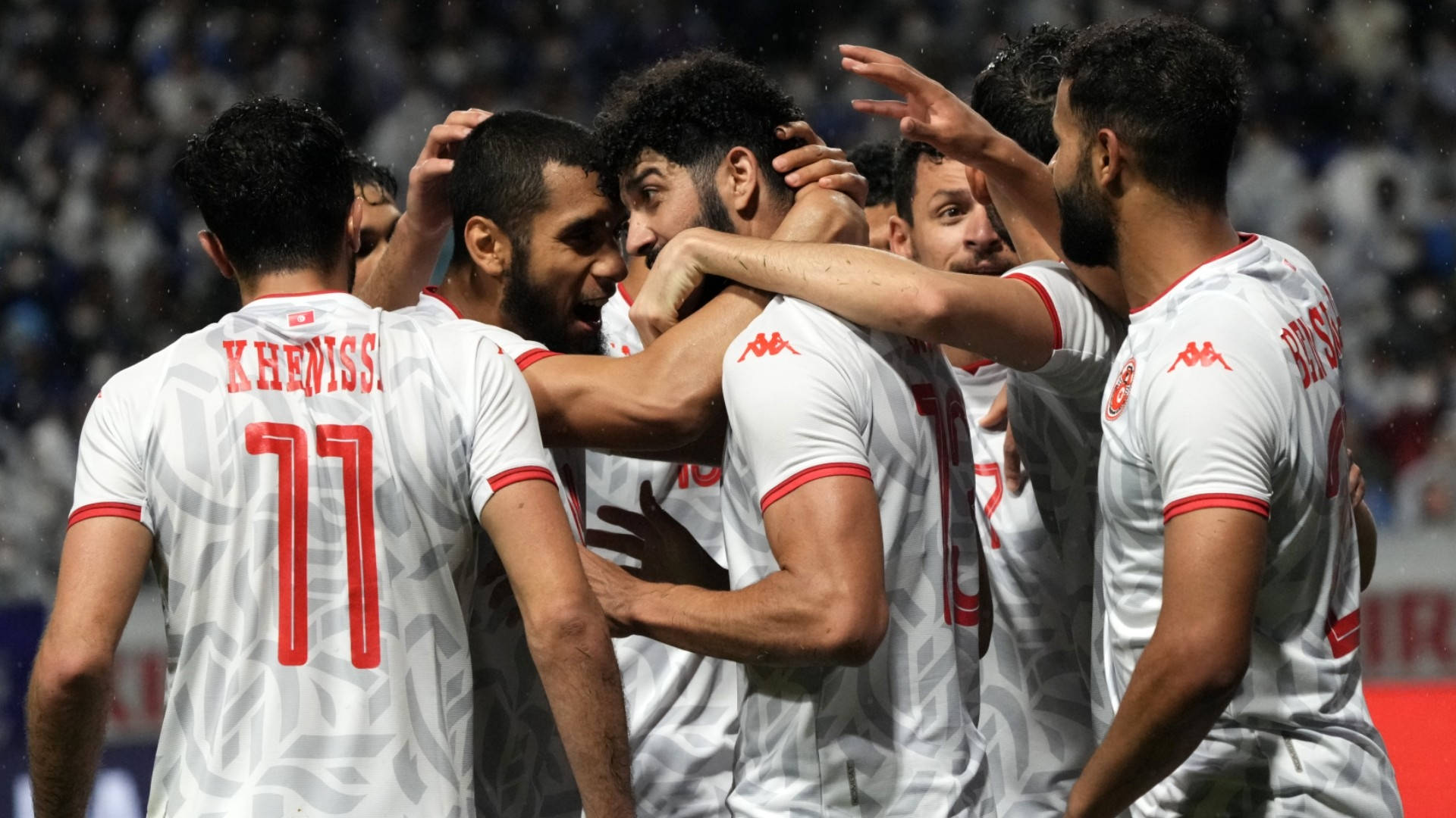 Tunisia 桌布 壁紙 — 身穿白色球衣帶紅色點綴的足球運動員在球場燈光下緊緊擁抱慶祝入球。