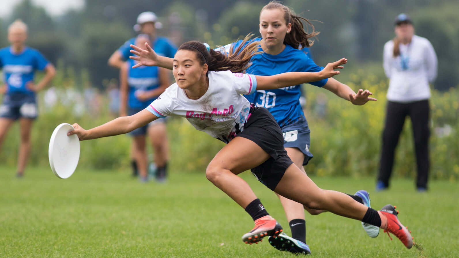 身穿白色球衣的 Ultimate Frisbee 女子運動員 1080p 背景 壁紙 — 一名女運動員向前飛撲接住飛盤，而對手球員在草地上逼近。