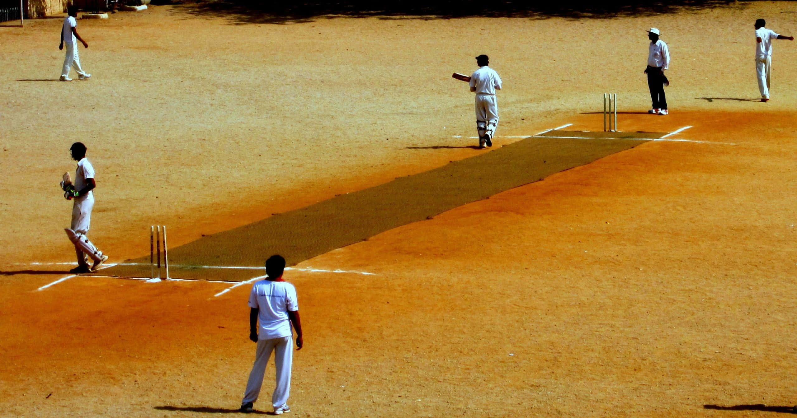 Cricket 桌布 壁紙 — 身穿白色球衣的板球運動員在塵土飛揚的橙色球場上，在明亮的日光下競爭，投下長長的影子。
