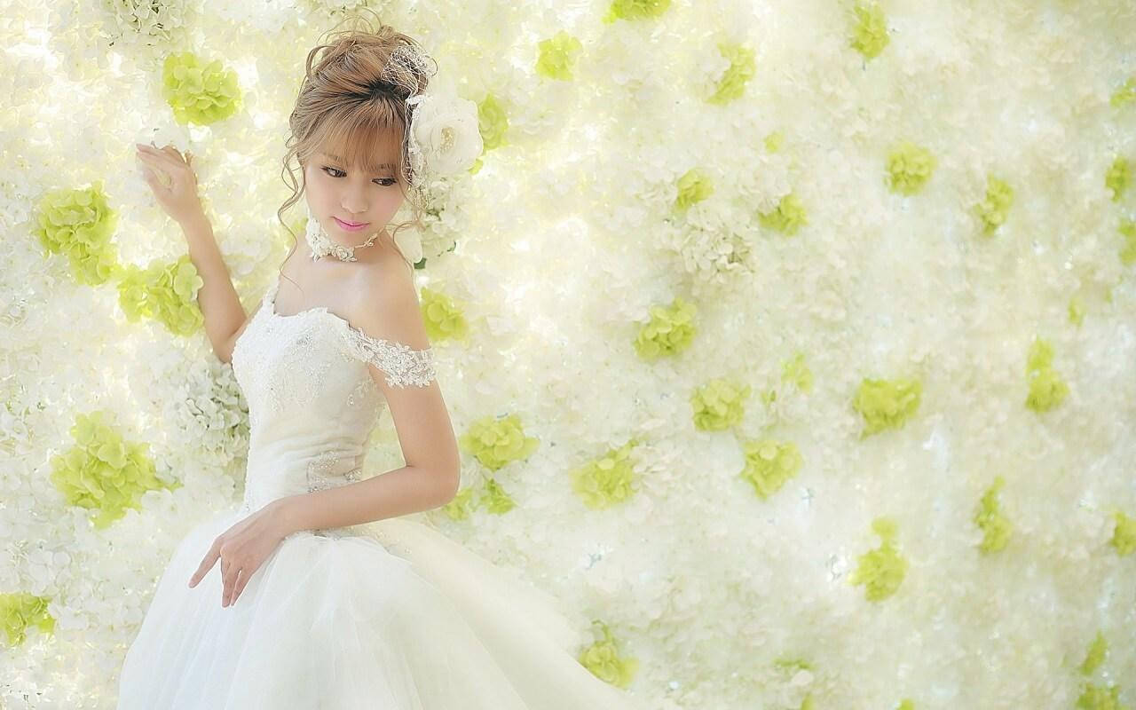 身穿白裙的可愛新娘 壁紙 — 一位身穿白色蕾絲婚紗的新娘，在白色和淺綠色繡球花牆前優雅地擺姿勢。