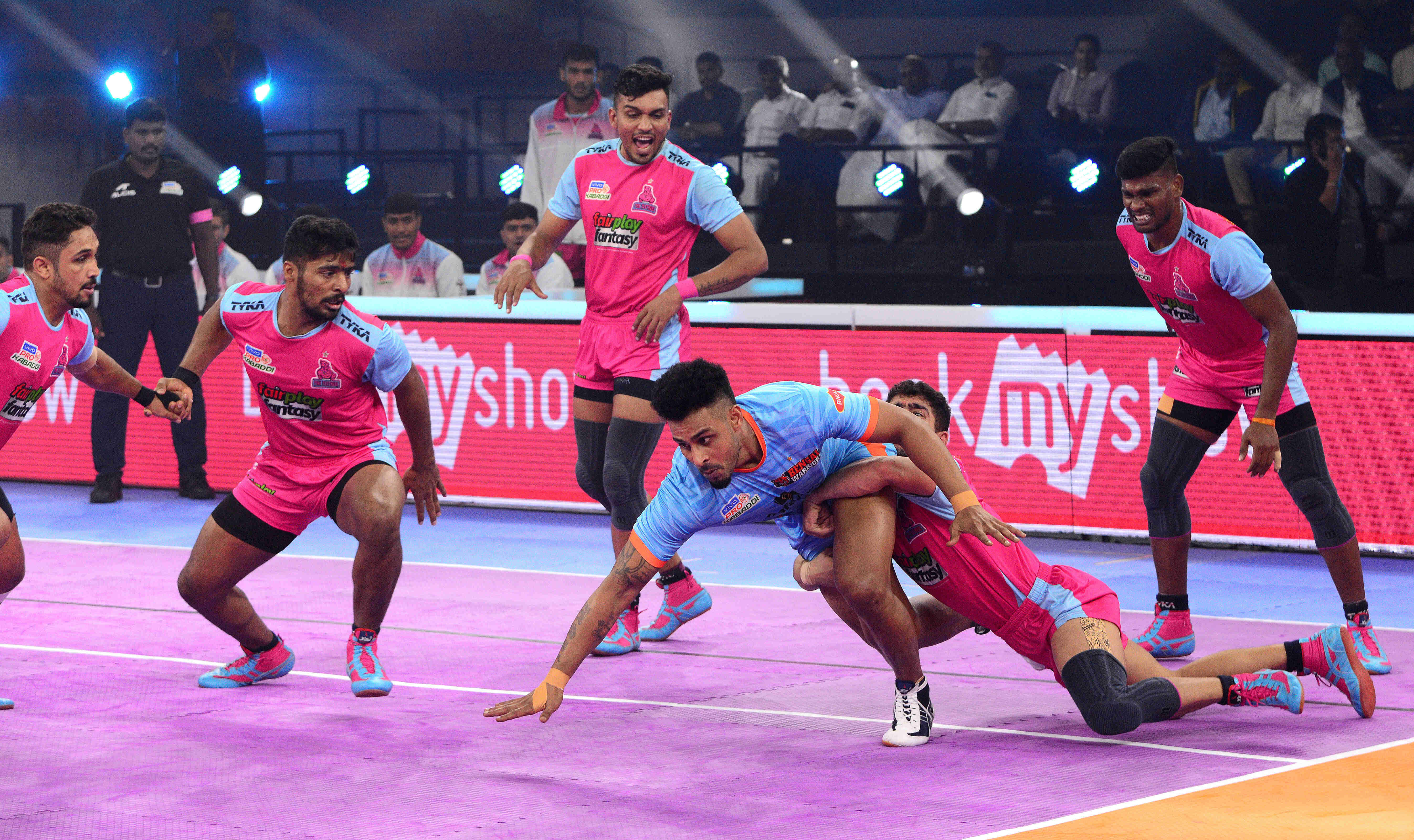 Kabaddi 桌布 壁紙 — 身穿粉紅色和藍色球衣的運動員在明亮的場館燈光下，在充滿活力的紫色球場上激烈競爭。