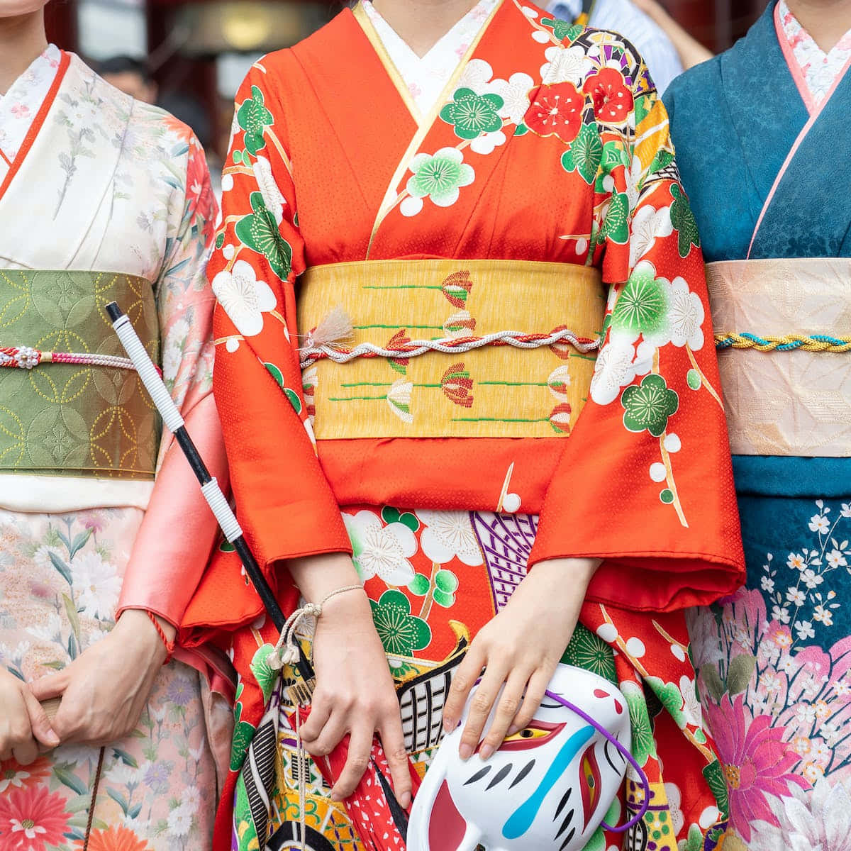 身穿粉紫和服的年輕亞洲女性 壁紙 — 三位身穿鮮豔傳統和服、帶有花卉圖案嘅女士企埋一齊，手持裝飾面具。