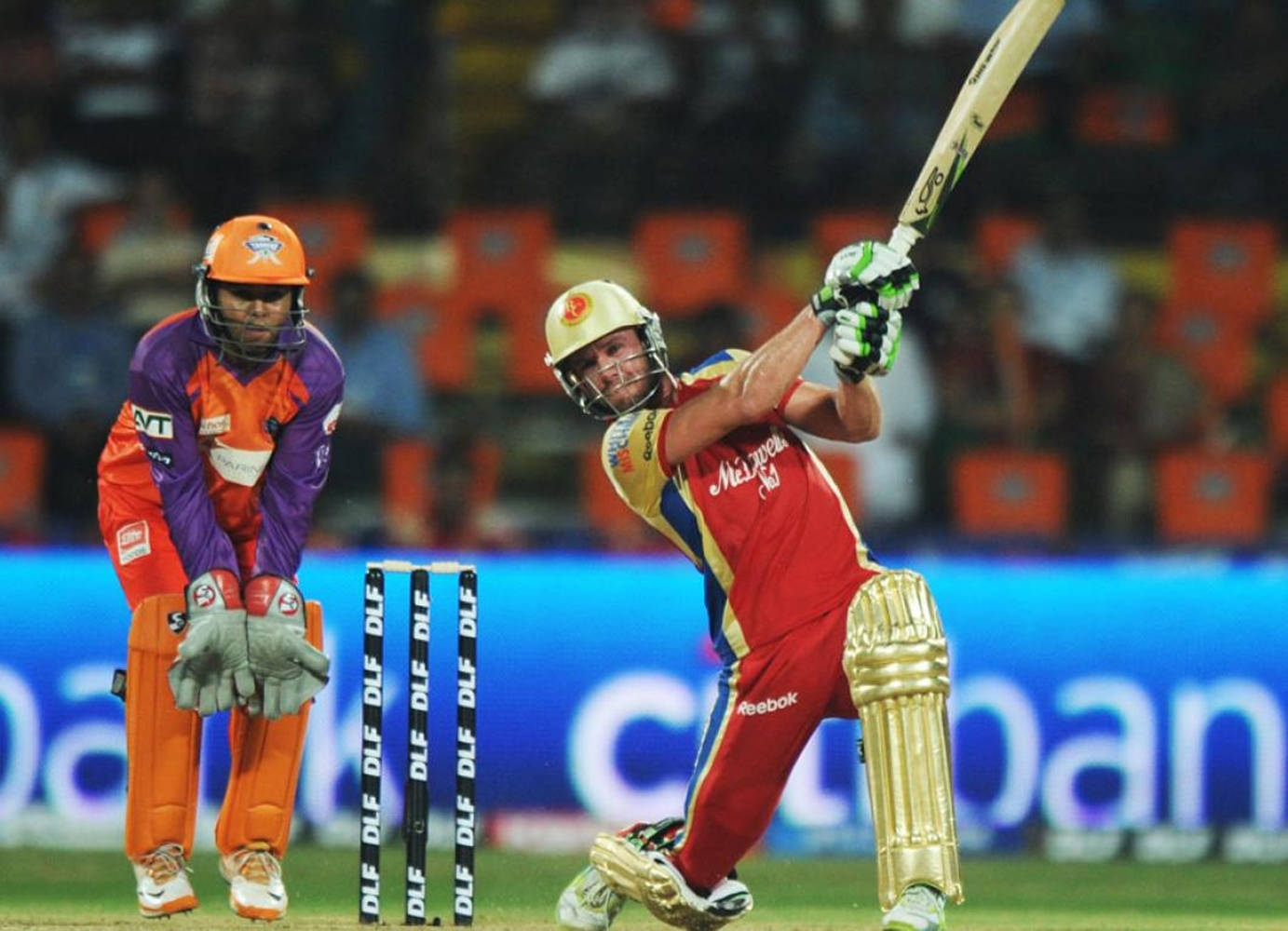 AB De Villiers RCB IPL 2011 壁紙 — 身穿紅色球衣的板球擊球手在球場燈光下強力揮棒，而身穿橙色裝備的守門員則蹲在門柱後面。