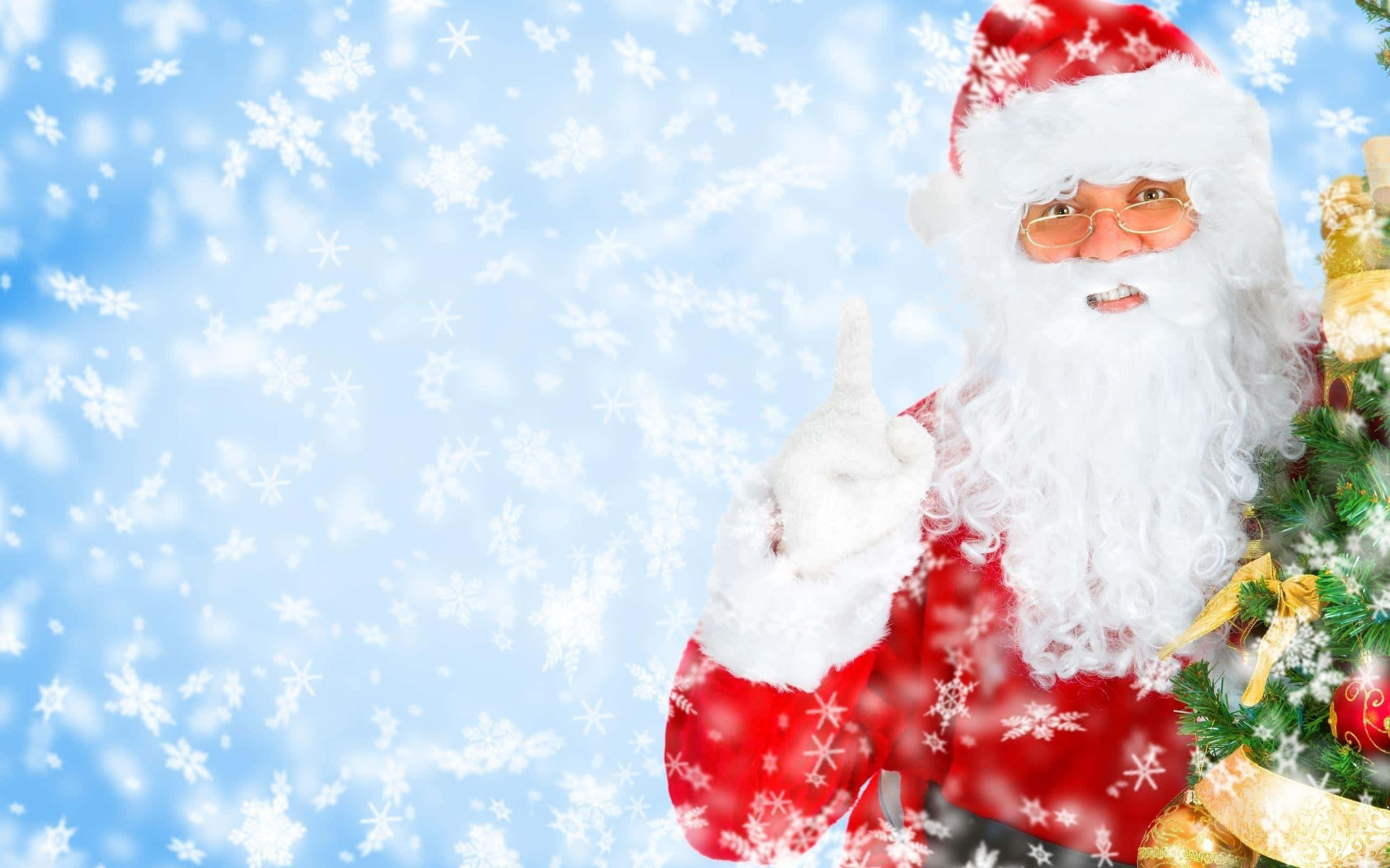 Santa Claus 桌布 壁紙 — 身穿紅色西裝的聖誕老人，在飄雪藍天和裝飾聖誕樹的背景下向上指。