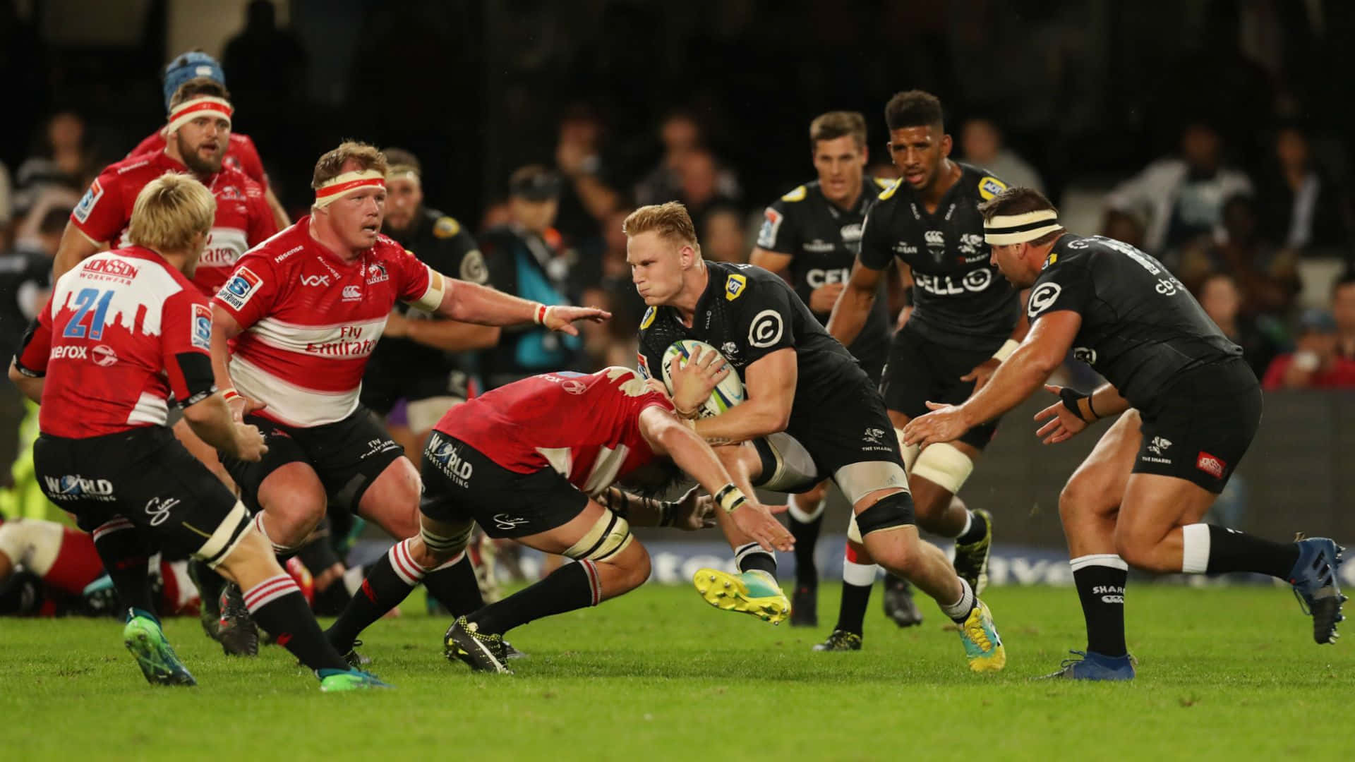 Rugby 桌布 壁紙 — 身穿紅黑色球衣的欖球運動員在體育場燈光下，於充滿活力的綠色場地上激烈碰撞。