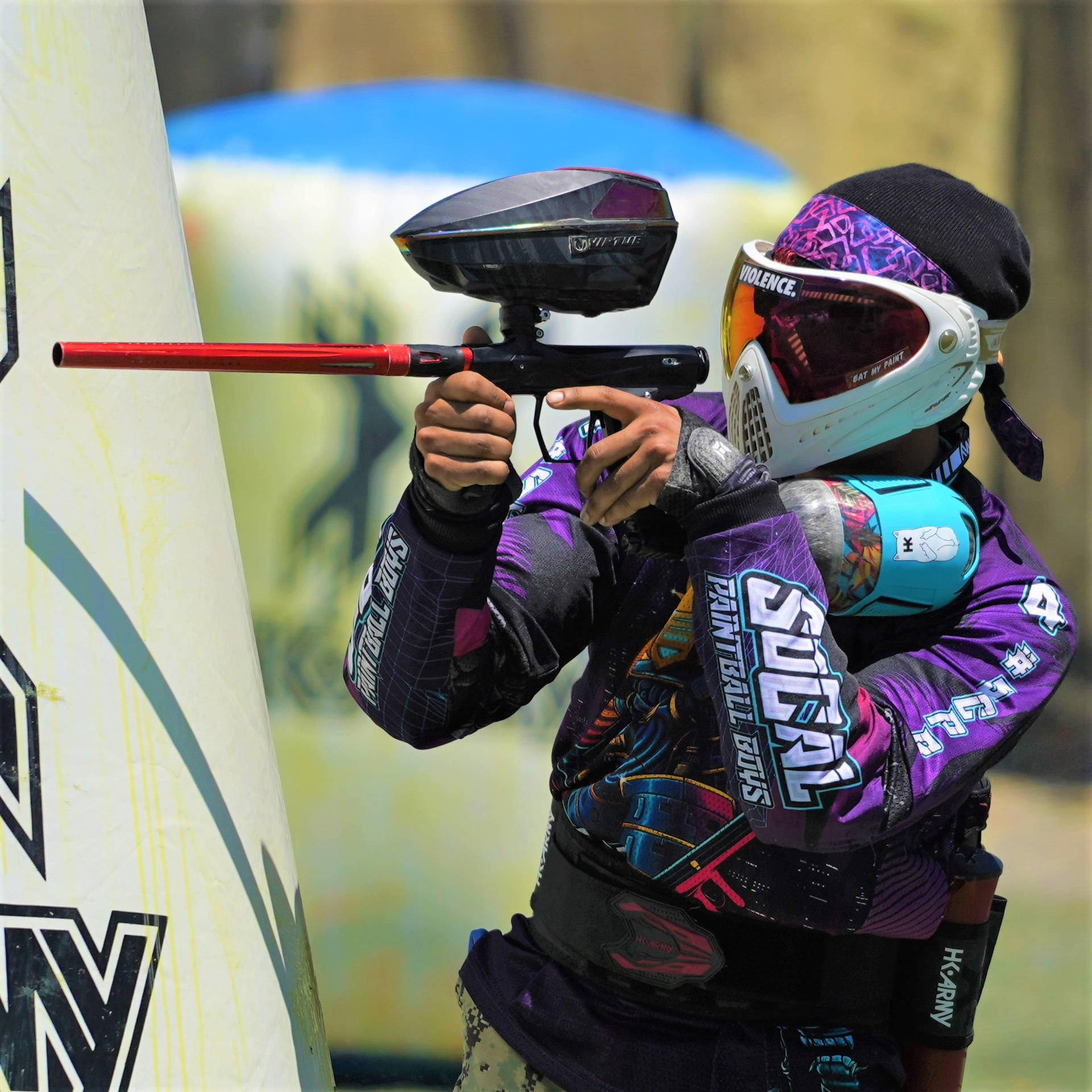 身穿紫色「Socal Paintball Boys」球衣的彩彈玩家 壁紙 — 一名身穿鮮紫色裝備的彩彈玩家在明亮的日光下，於白色掩體後方瞄準一支紅色槍管的標記器。