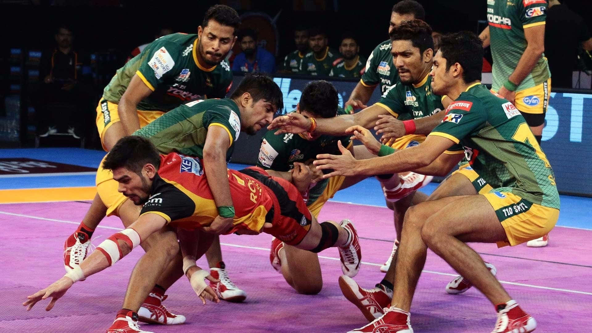 Kabaddi 桌布 壁紙 — 身穿綠色、黃色和紅色制服嘅激烈卡巴迪選手，喺明亮嘅場館燈光下，喺充滿活力嘅紫色場地上激烈搏鬥。