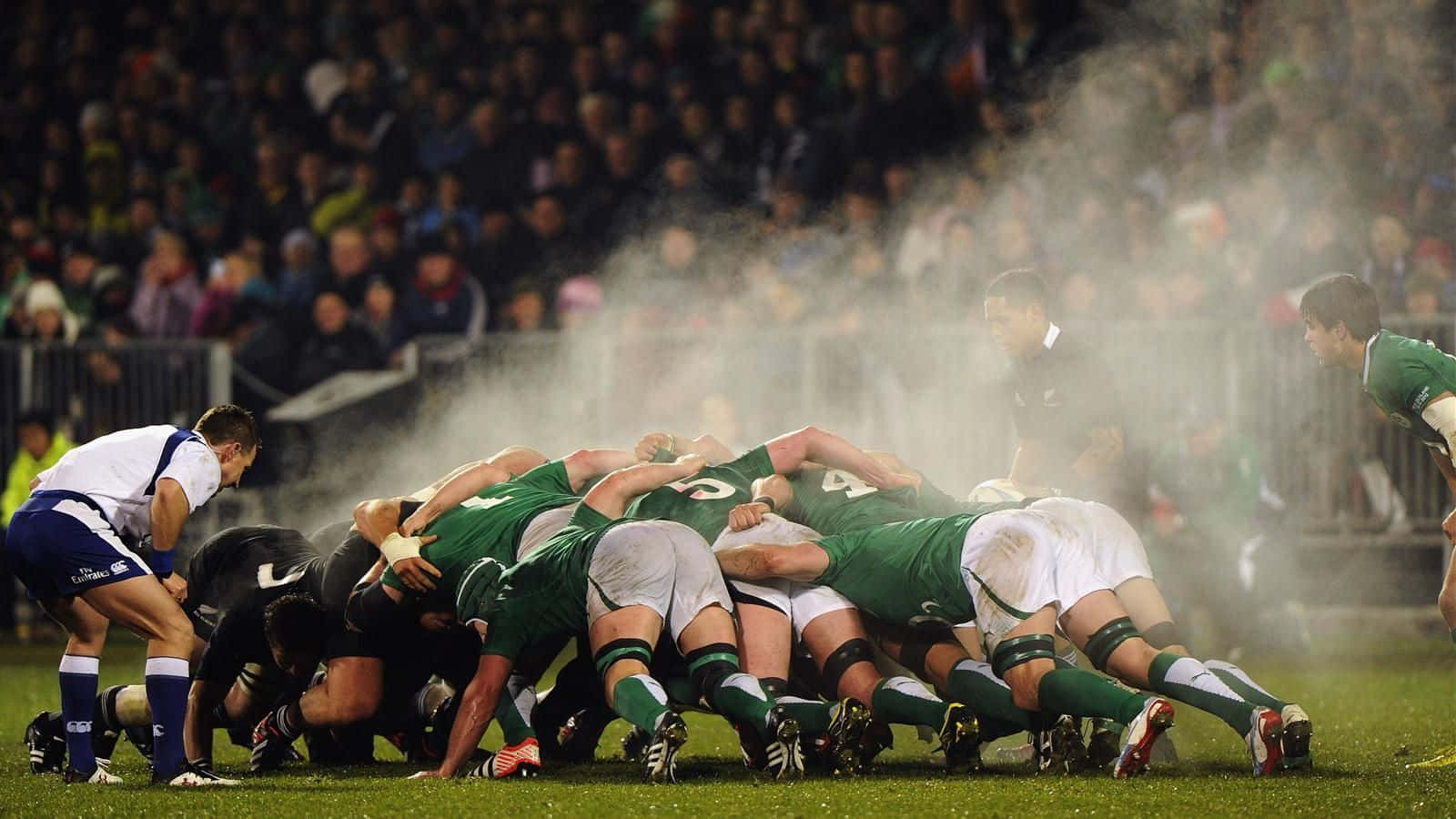 Rugby 桌布 壁紙 — 身穿綠色和白色球衣的欖球運動員在草地上形成強大的爭球，霧氣在球場燈光下在他們周圍升起。