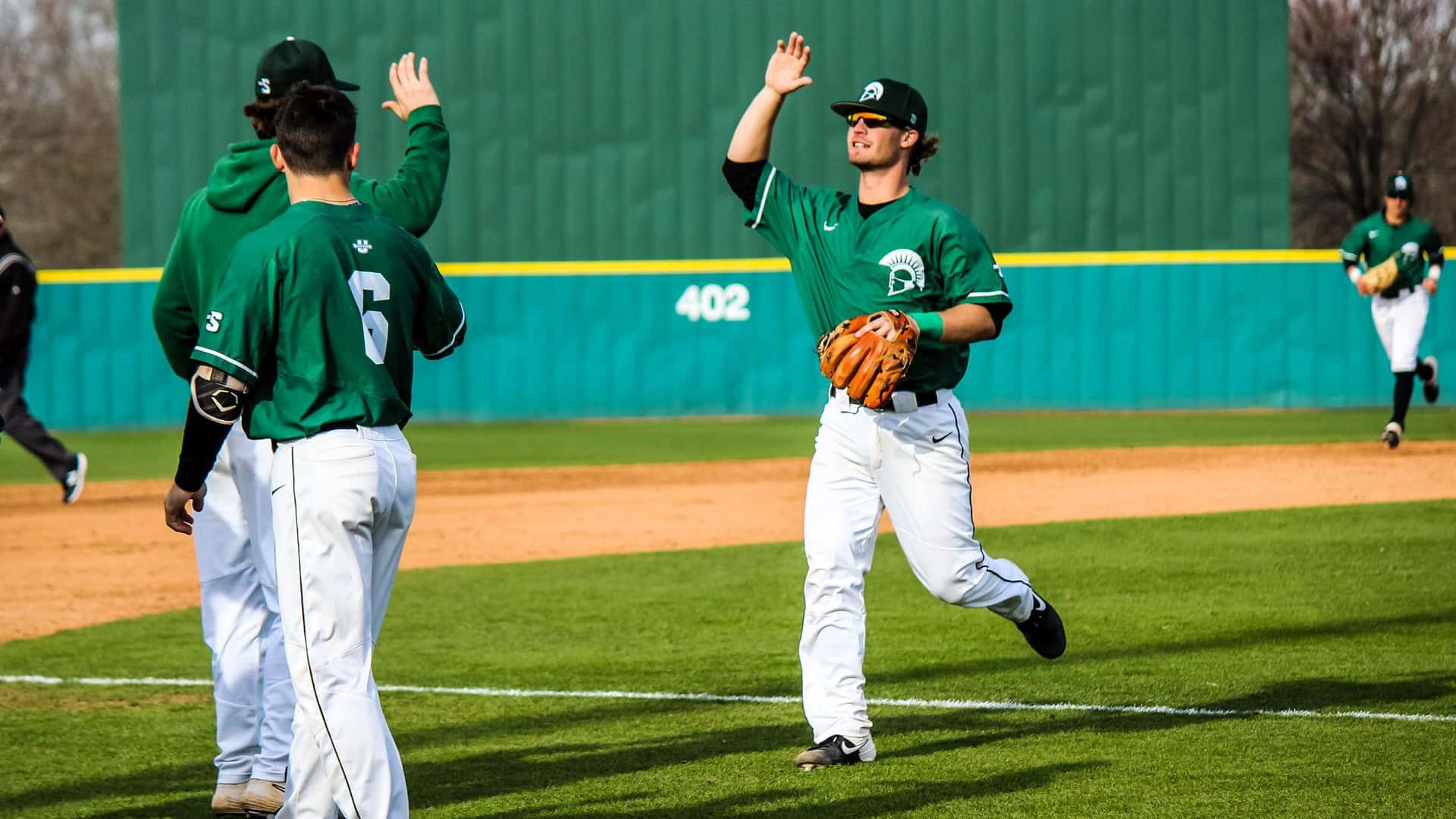 College Baseball 桌布 壁紙 — 身穿綠色球衣的棒球運動員在陽光明媚的球場上慶祝，背景有青綠色牆壁和數字402。