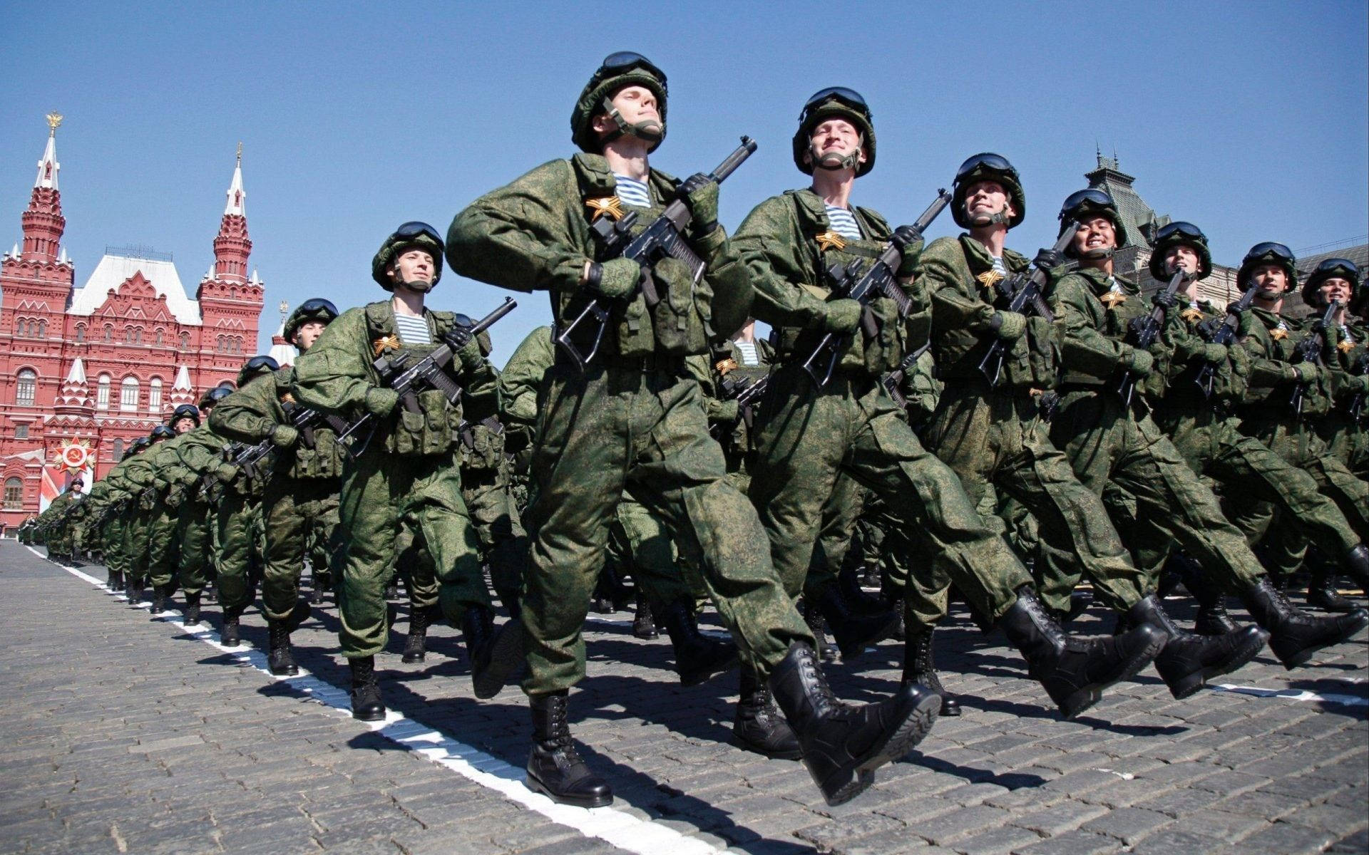 Russia 桌布 壁紙 — 身穿綠色迷彩軍服的士兵在晴朗的藍天下，以精確的隊形穿過鵝卵石廣場，背景是歷史悠久的紅色建築。