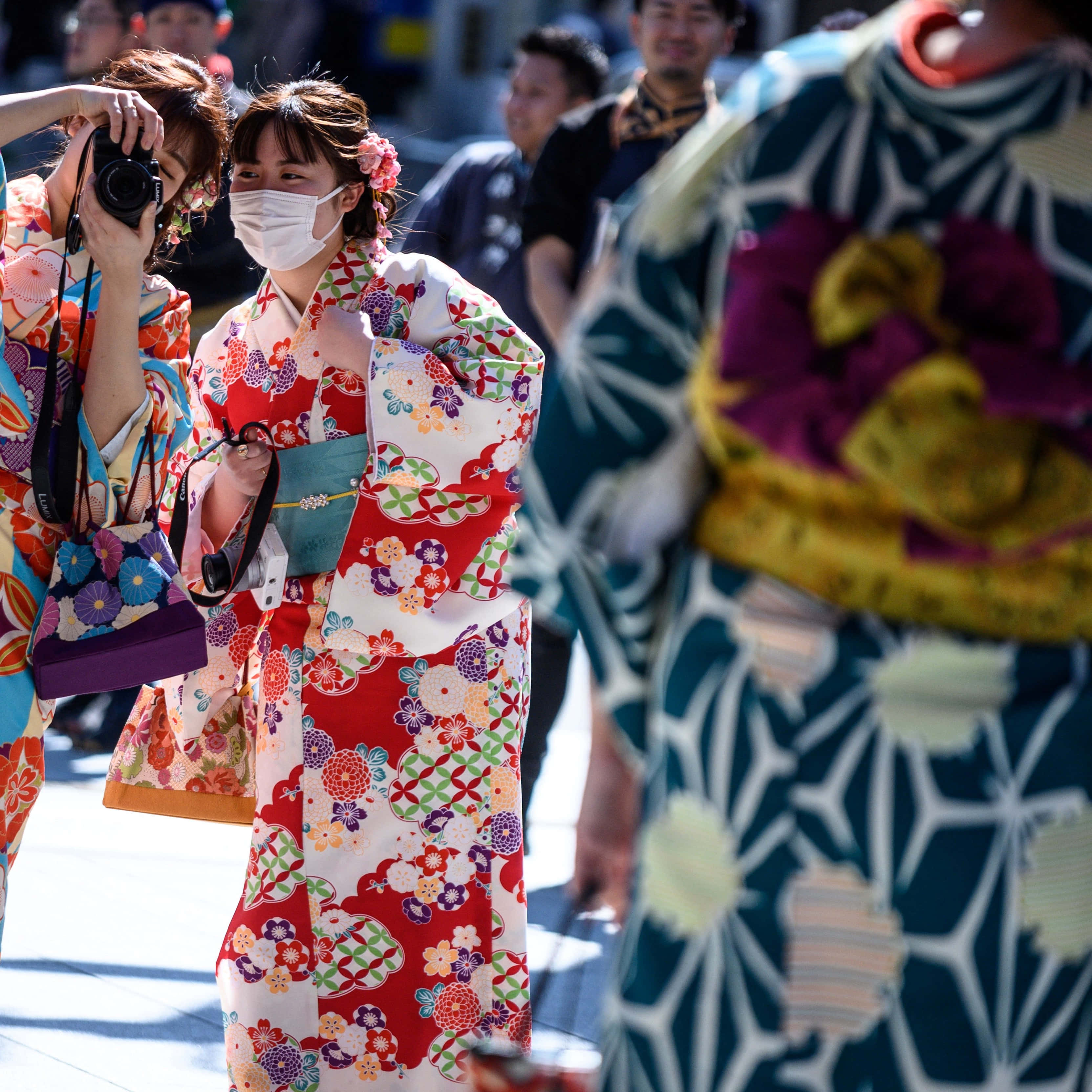 身穿色彩繽紛和服的日本女性 壁紙 — 一位年輕女子身穿鮮豔的紅色花卉和服，戴著白色口罩，走過繁忙的街道。