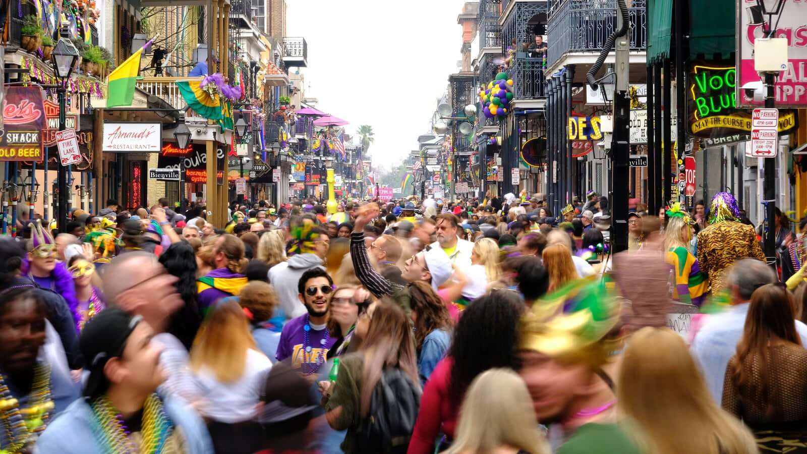 Mardi Gras 桌布 壁紙 — 身穿色彩繽紛狂歡節服飾和珠鏈的狂歡人群，擠滿了一條充滿節日氣氛的街道，兩旁是裝飾華麗的建築物和懸掛的旗幟。