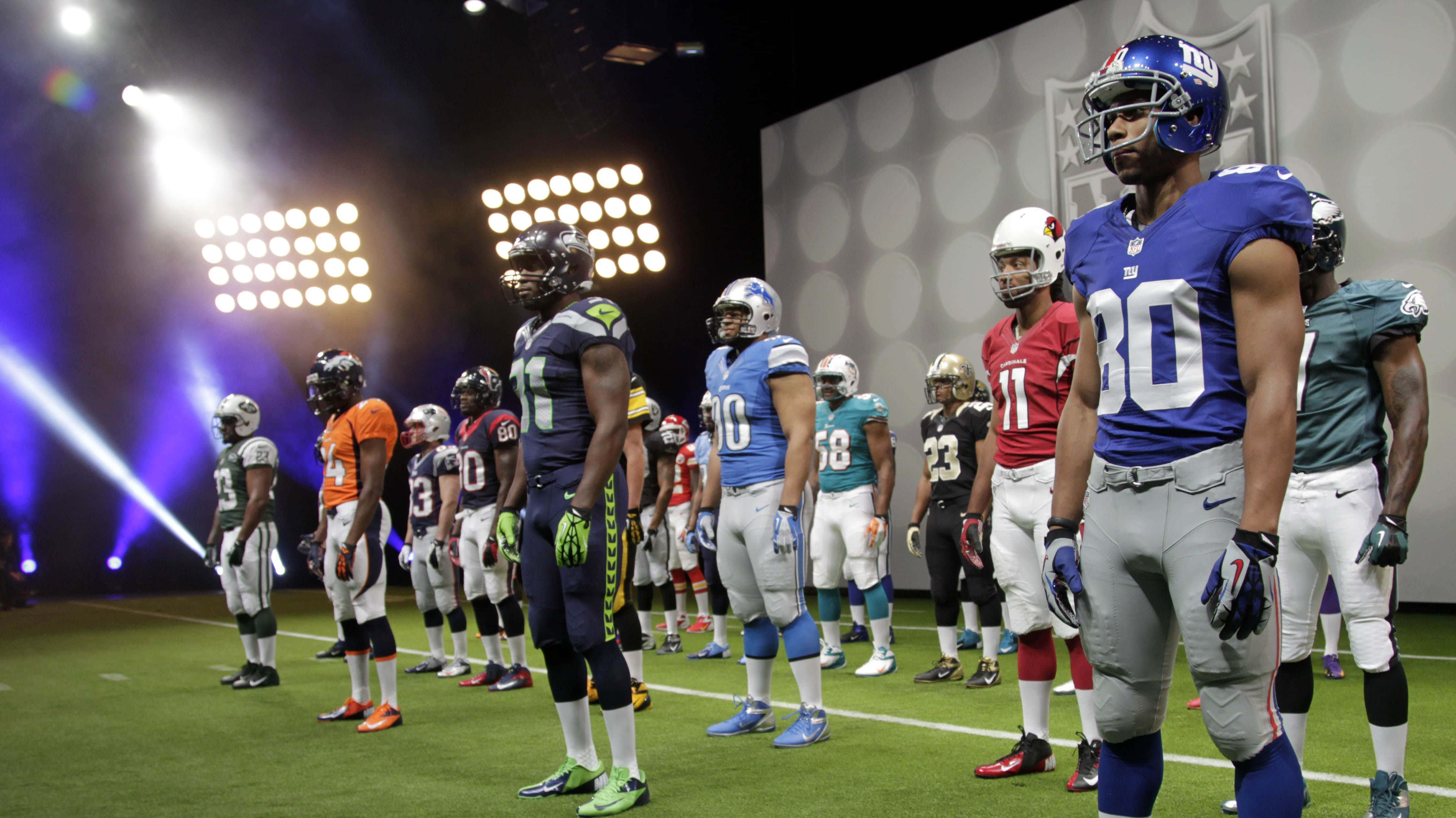 NFL 桌布 壁紙 — 身穿色彩繽紛球衣的NFL美式足球員在球場燈光下排成一線，站在明亮的場地上。