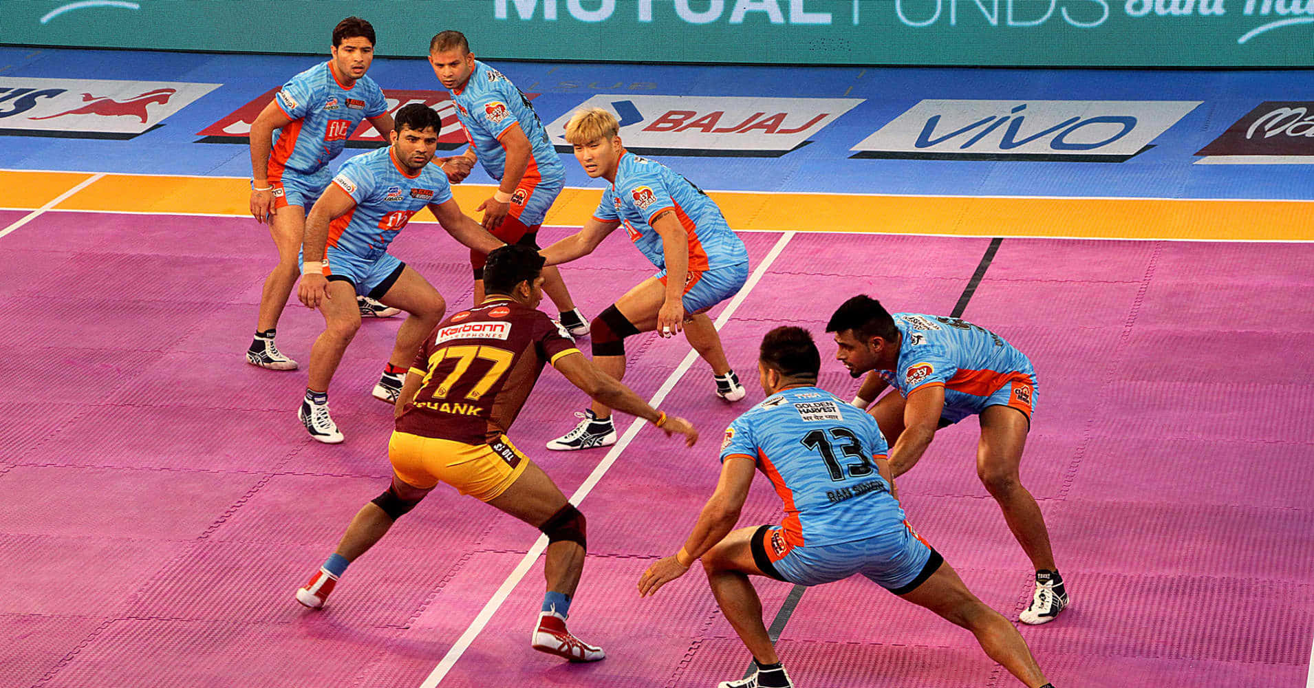Kabaddi 桌布 壁紙 — 身穿藍色和栗色球衣的球員在充滿活力的粉紅色室內球場上激烈爭球。
