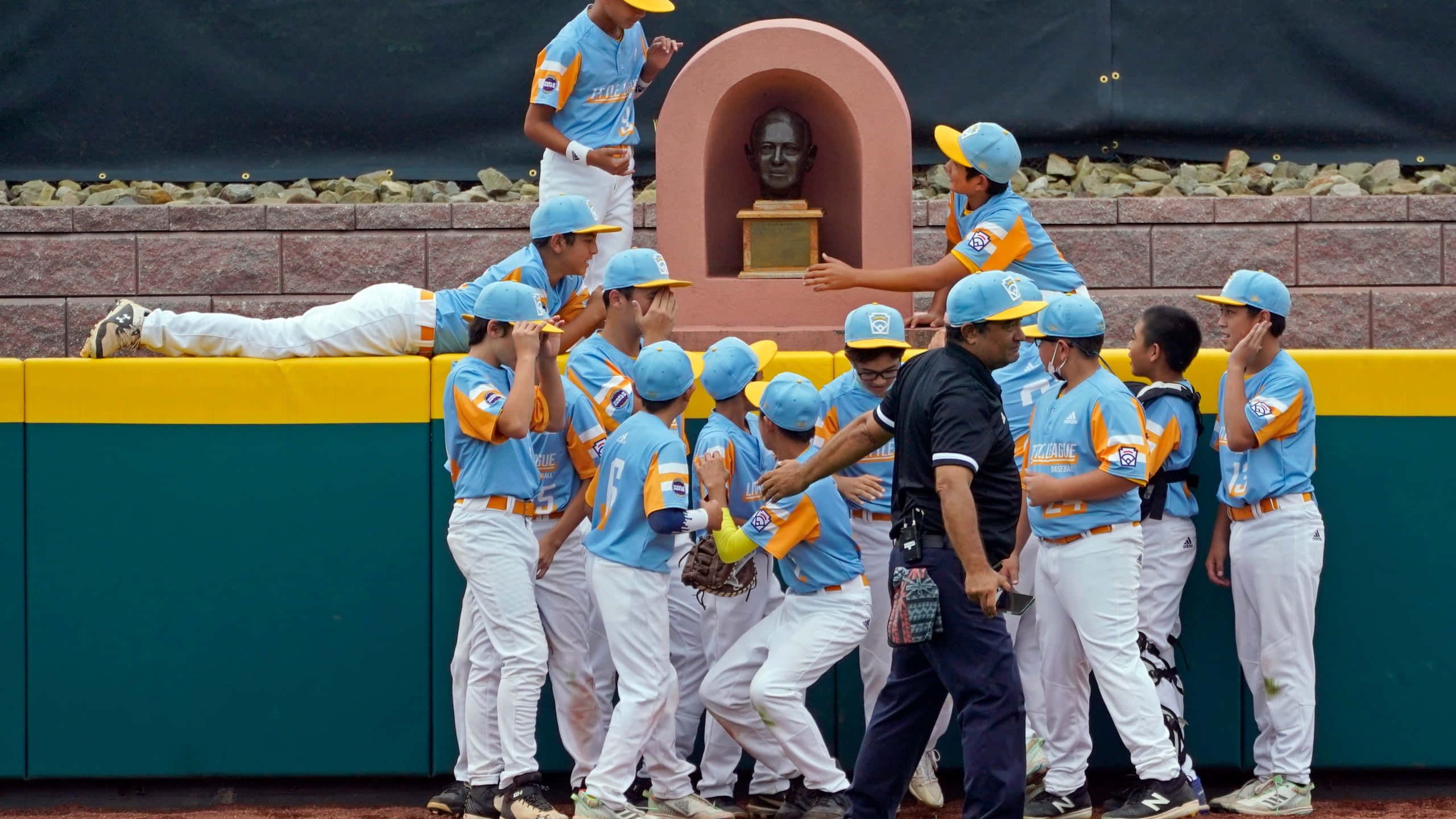 Little League Baseball 桌布 壁紙 — 身穿藍色和橙色球衣的年輕棒球運動員在身後有雕像壁龕的球員休息區牆壁附近熱情慶祝。