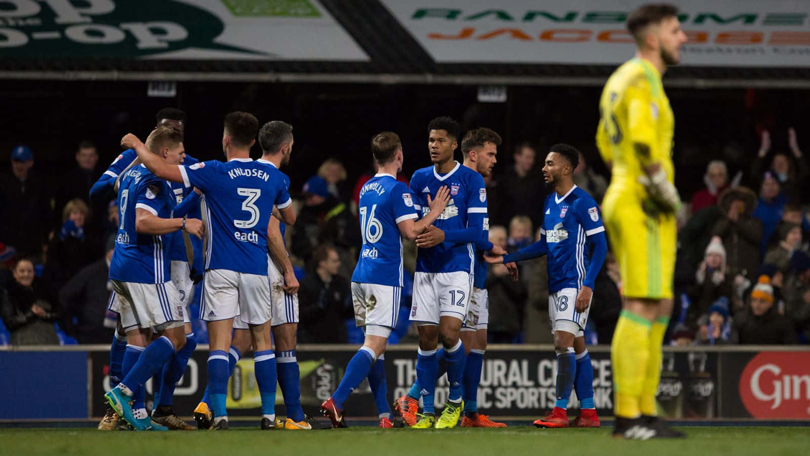 Ipswich Town 桌布 壁紙 — 身穿藍色球衣的足球運動員在燈光璀璨的球場上慶祝入球，附近有一位身穿黃色球衣的守門員。