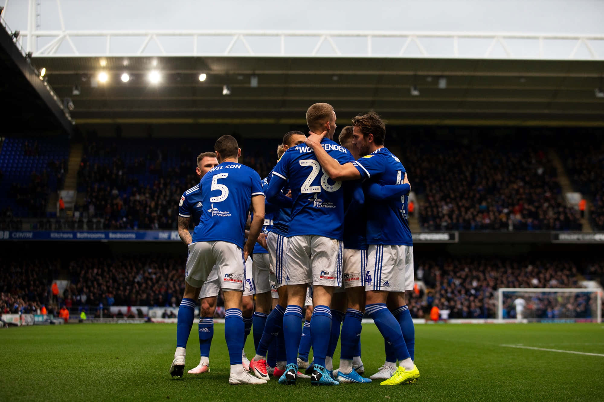 Ipswich Town 桌布 壁紙 — 身穿藍色球衣的足球運動員在球場燈光下的綠色草地上聚在一起，看台上有球迷。