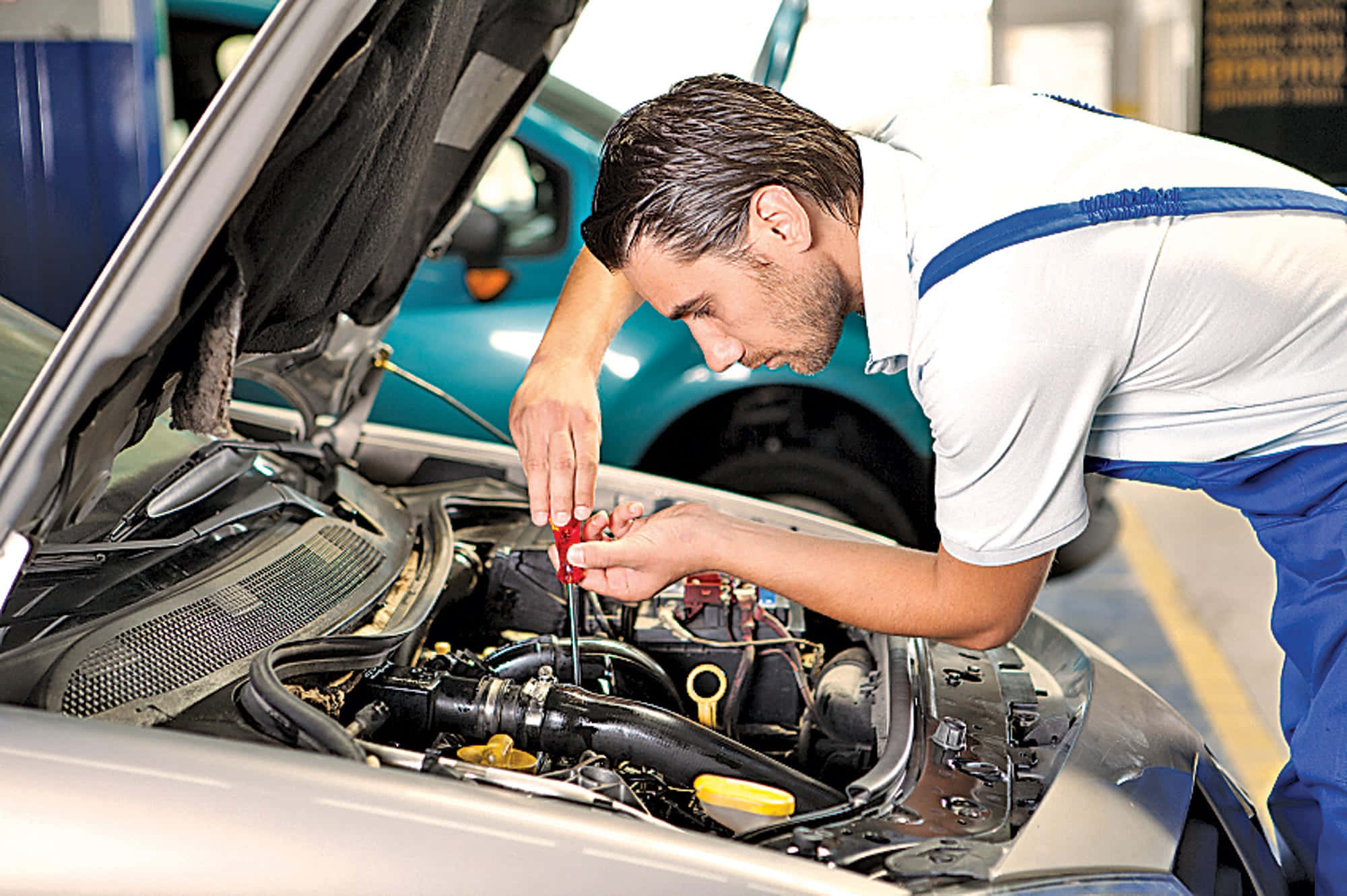 Car Repair 桌布 壁紙 — 身穿藍色連身工作服的機械師在車庫中，用紅色螺絲批調整銀色汽車打開的引擎蓋下的引擎部件。