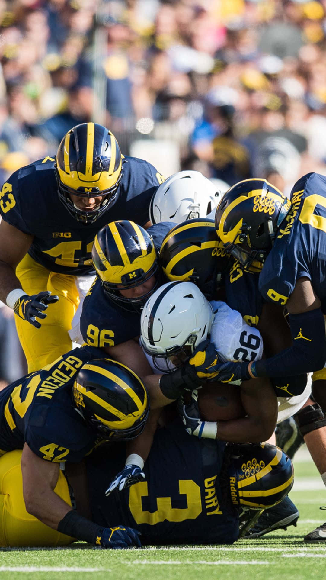 Michigan Wolverines 桌布 壁紙 — 身穿藍黃色球衣的足球員在日間比賽中於綠色球場上攔截對手。