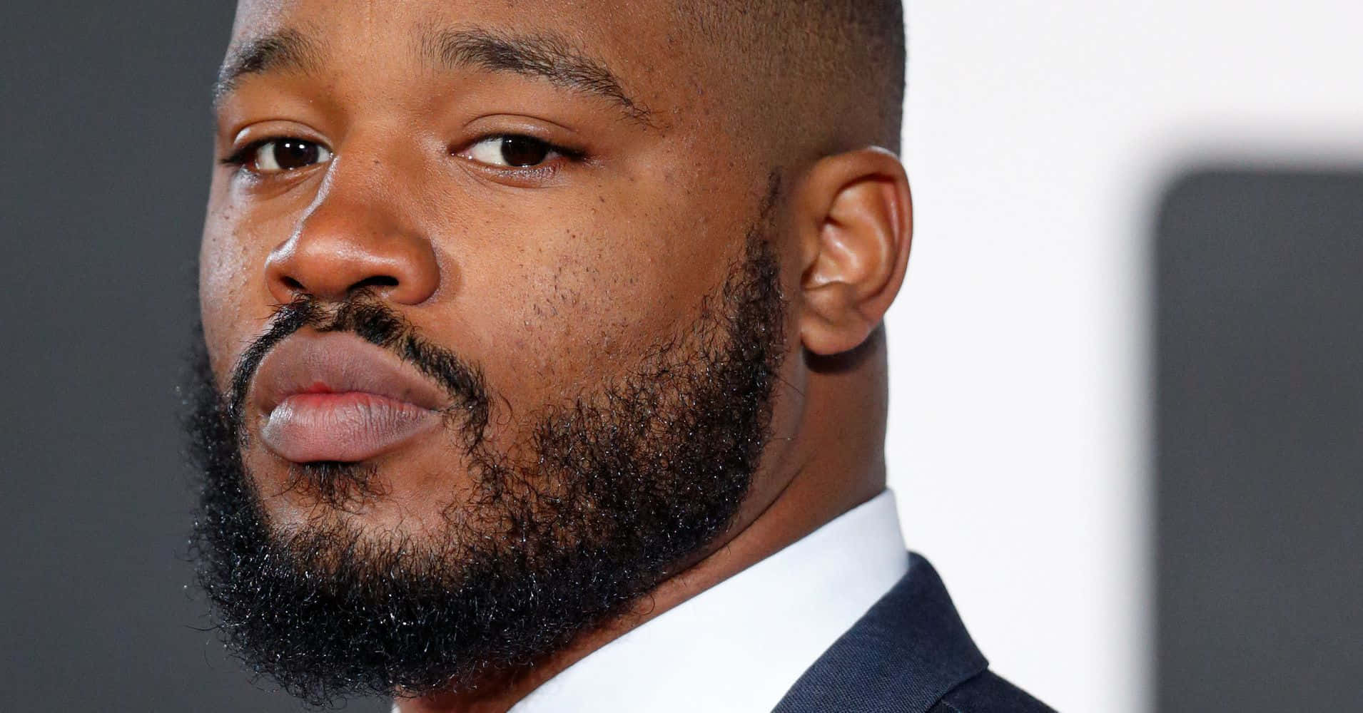 Ryan Coogler 桌布 壁紙 — 身穿西裝、留著濃密鬍鬚的男士特寫肖像，背景為模糊的灰色和白色。