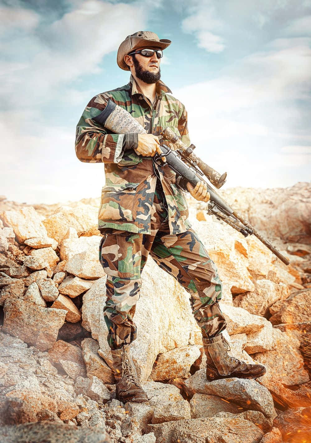 身穿軍裝持狙擊槍的男子 壁紙 — 一名留鬍鬚的士兵身穿迷彩裝備，站在岩石地形上，在明亮的天空下持步槍。