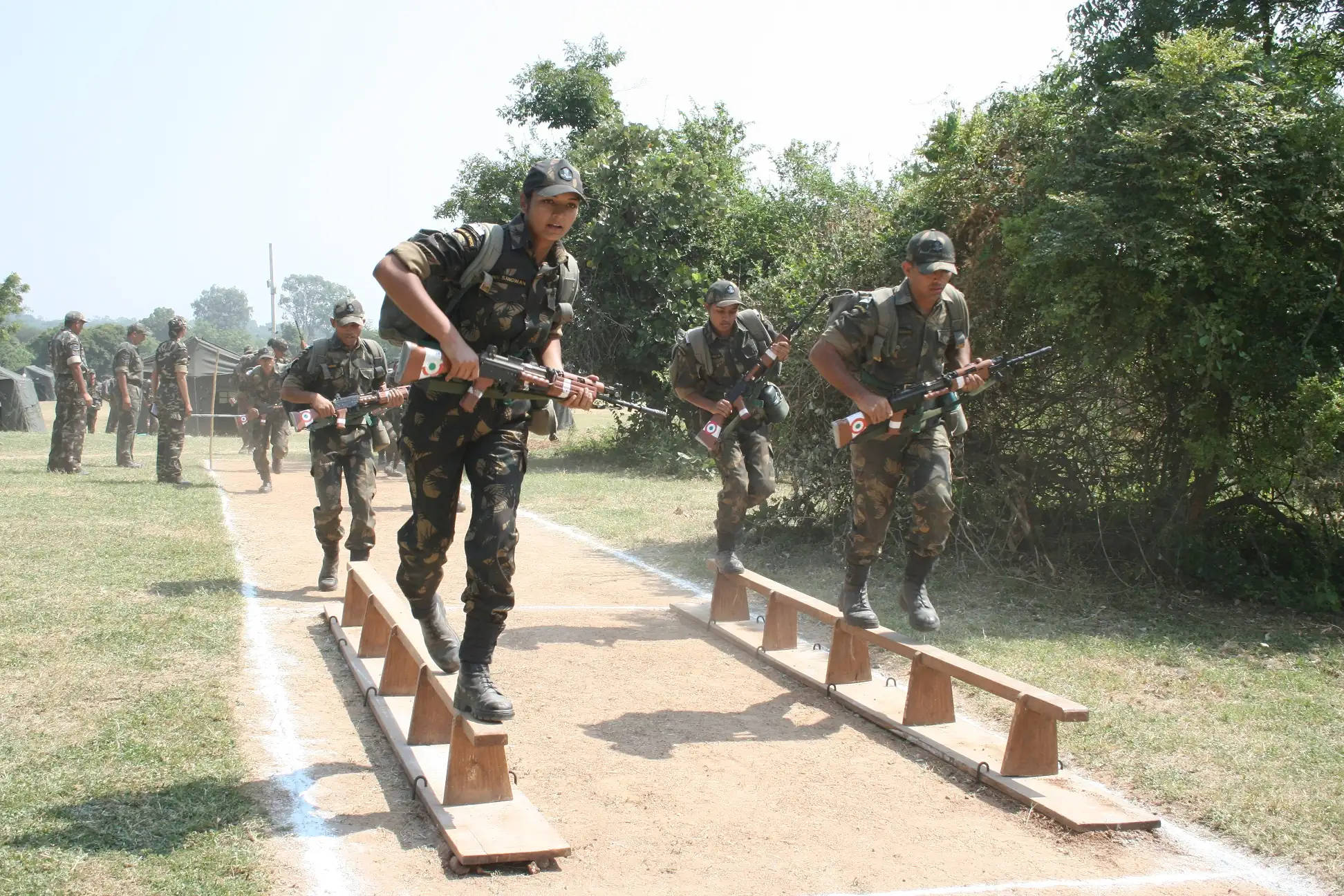 Indian Army 桌布 壁紙 — 身穿迷彩裝備的士兵在明亮的日光下穿越木製障礙訓練場，背景有樹木。