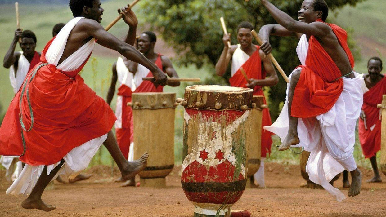 Africa 桌布 壁紙 — 身穿鮮豔紅白布料的舞者在明亮的日光下，在戶外泥地上充滿活力地表演大鼓。