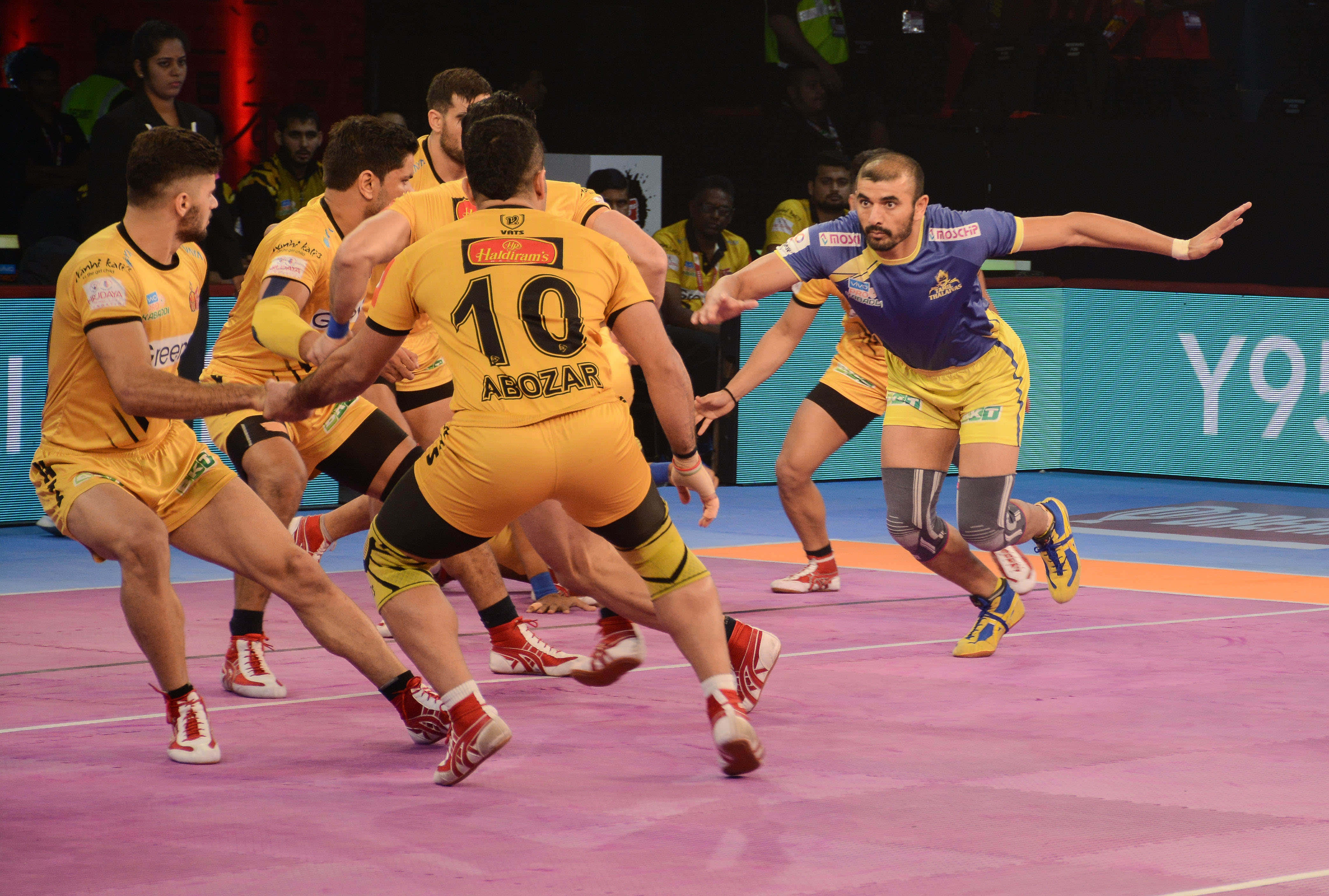 Kabaddi 桌布 壁紙 — 身穿黃色和藍色球衣的手球運動員在明亮的場館燈光下，在充滿活力的粉紅色球場上激烈競爭。