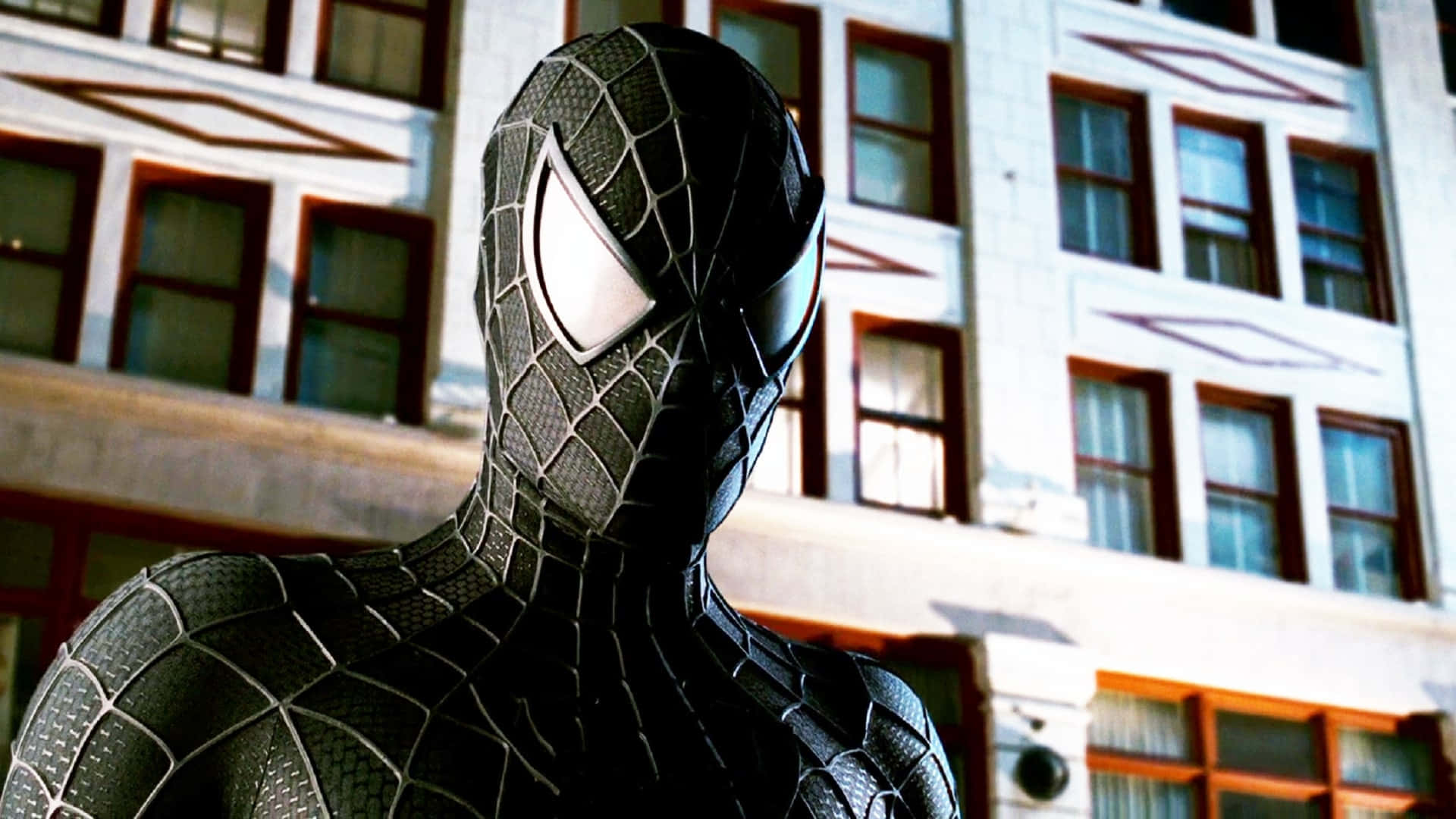 Spider Man 3 桌布 壁紙 — 身穿黑色戰衣的蜘蛛俠站在陽光普照的城市建築前，戰衣上有精細的網紋圖案，眼睛發出白光。