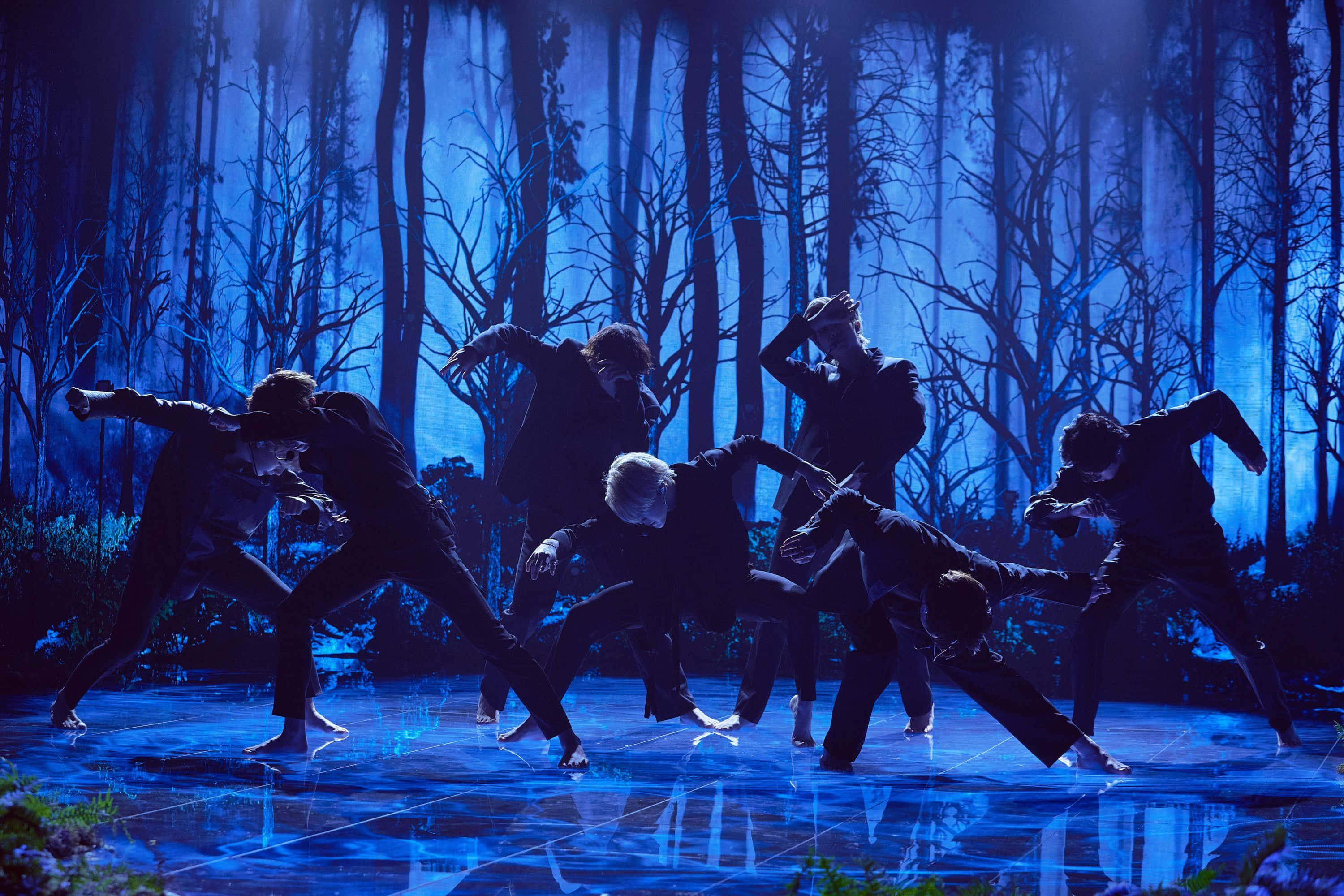Bts Performance 桌布 壁紙 — 身穿黑色西裝的舞者在發光的藍色舞台上，以迷霧森林為背景，擺出動態姿勢。