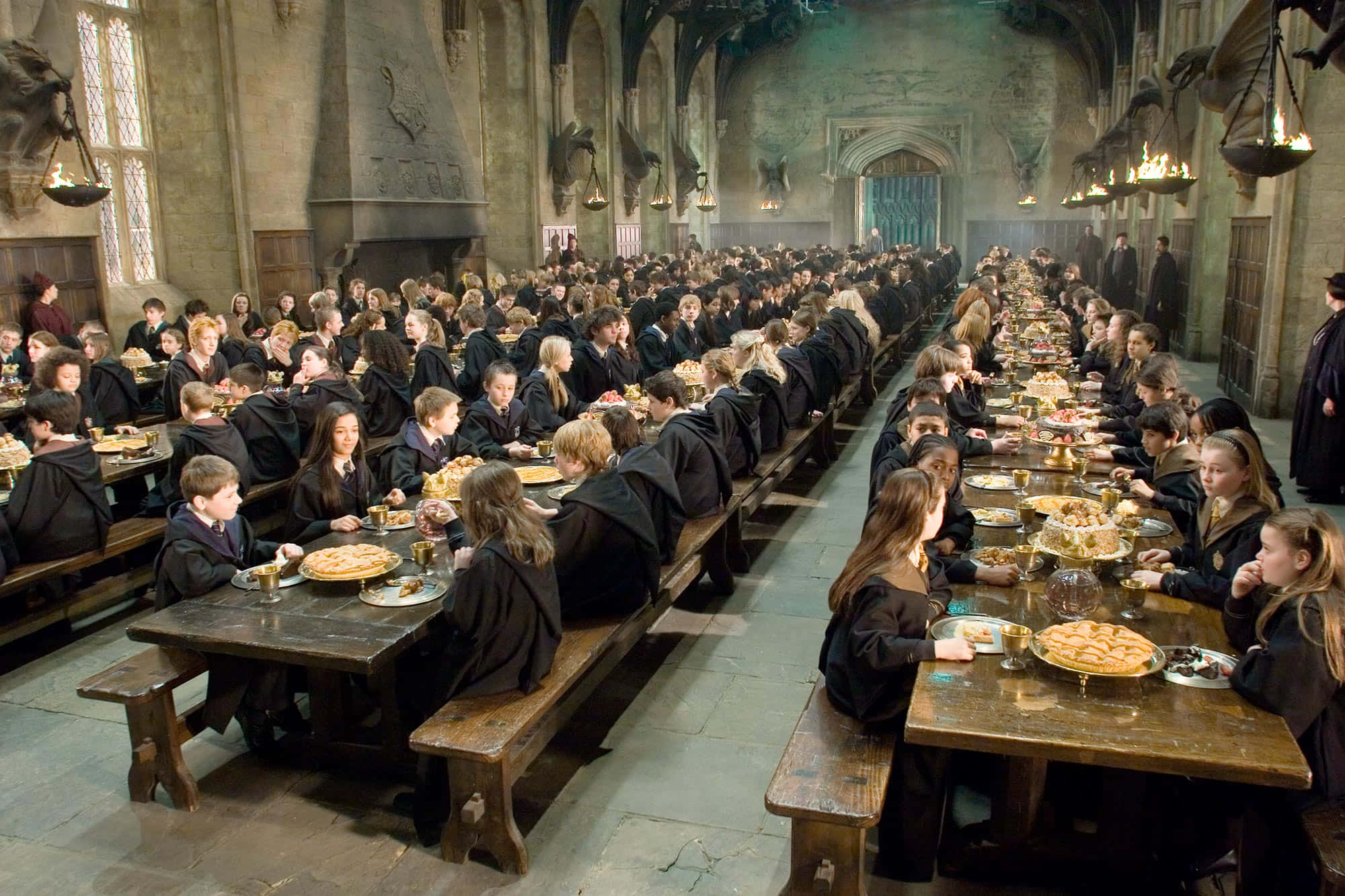 The Hogwarts Great Hall 桌布 壁紙 — 身穿黑色長袍的學生在高天花和掛著火把的宏偉石廳中的長木桌上用餐。