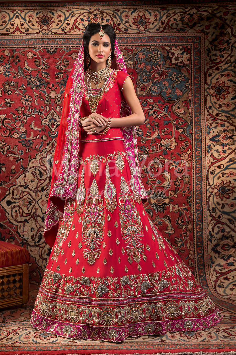 身著傳統婚紗的印度新娘 壁紙 — 一位身穿鮮紅色刺繡 lehenga 並飾有金色細節的女性，優雅地站在華麗圖案地毯前。