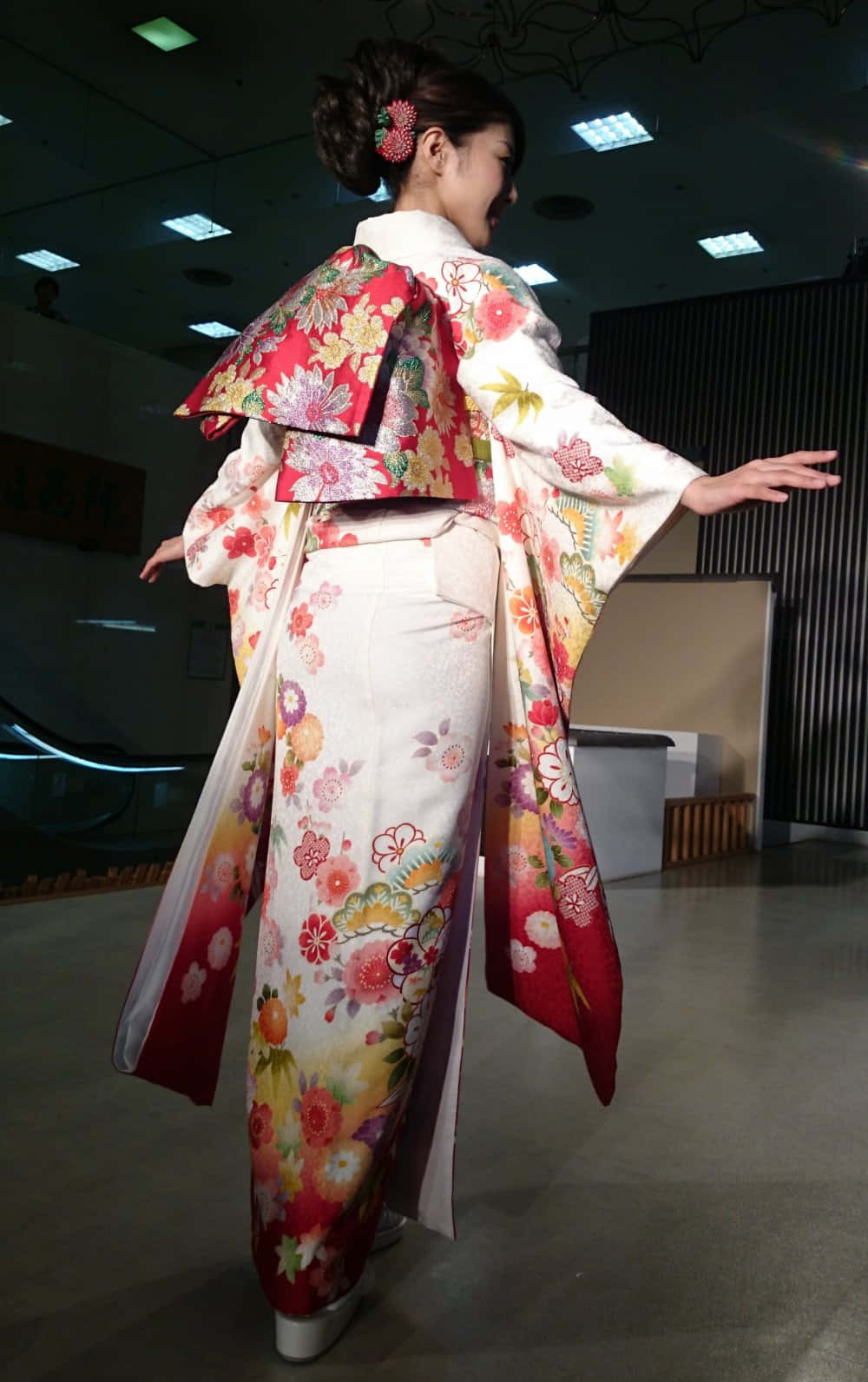 身著傳統日本和服的女子於庭園中 壁紙 — 一位身穿鮮豔花卉圖案和服、帶有紅色點綴的女性，在室內優雅地擺姿勢，背對鏡頭。