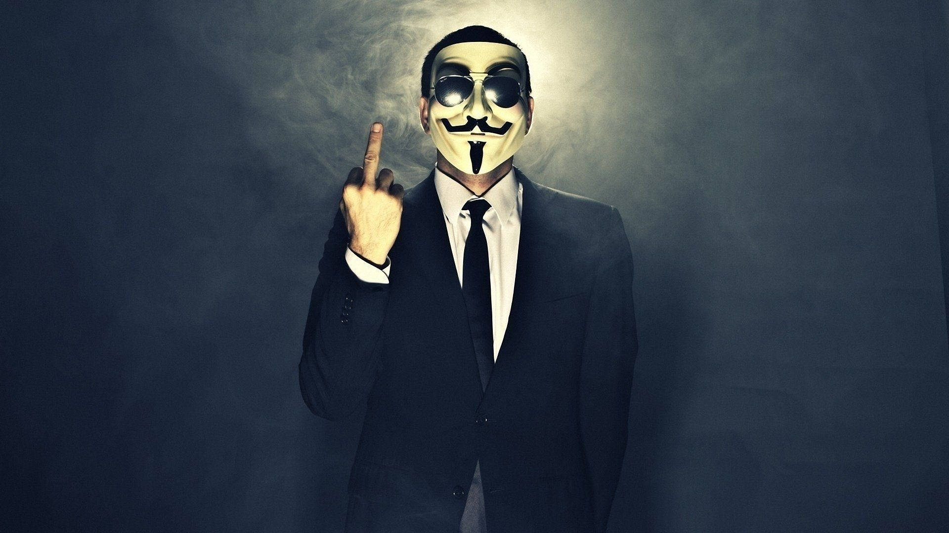 身著匿名西裝的神秘人物 壁紙 — 一個穿著西裝和蓋伊·福克斯面具的人，在煙霧瀰漫的黑暗背景下挑釁地豎起中指。