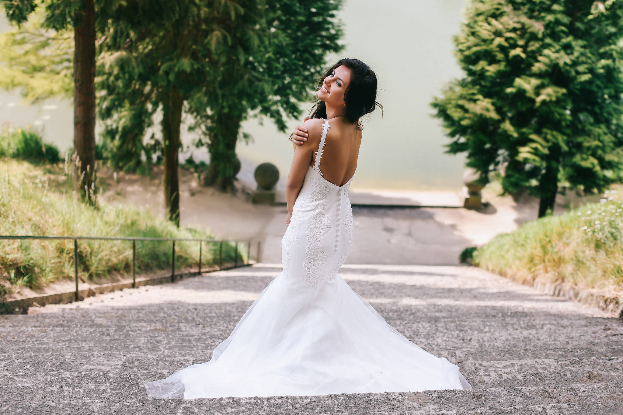 身著奢華婚紗的燦爛新娘 壁紙 — 一位身穿白色蕾絲美人魚婚紗的新娘，在綠樹成蔭的石徑上回頭微笑。
