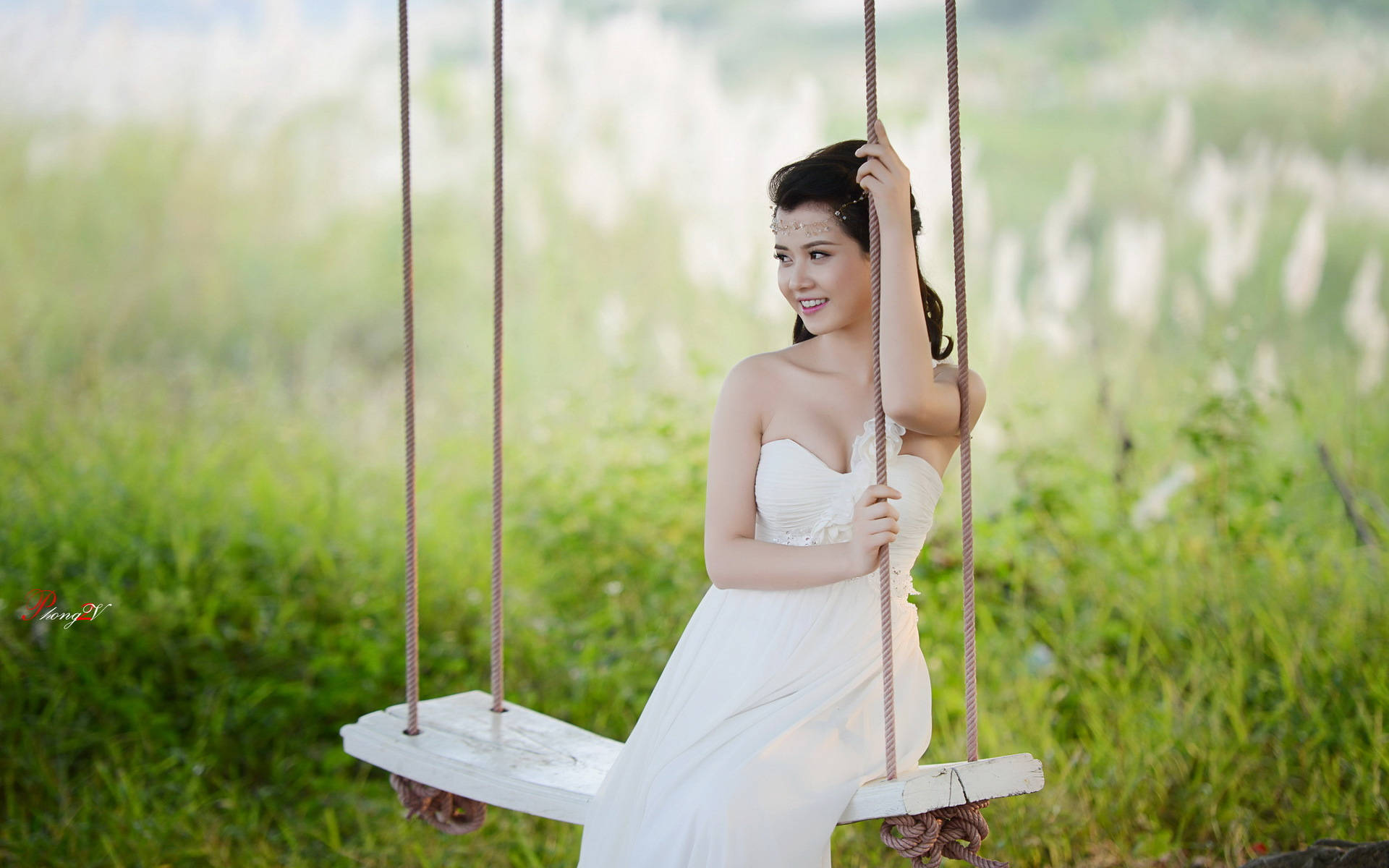 身著婚紗的燦爛新娘 壁紙 — 一位微笑的白裙女士優雅地坐在陽光普照的綠色田野中的木鞦韆上，背景有模糊的蘆葦。