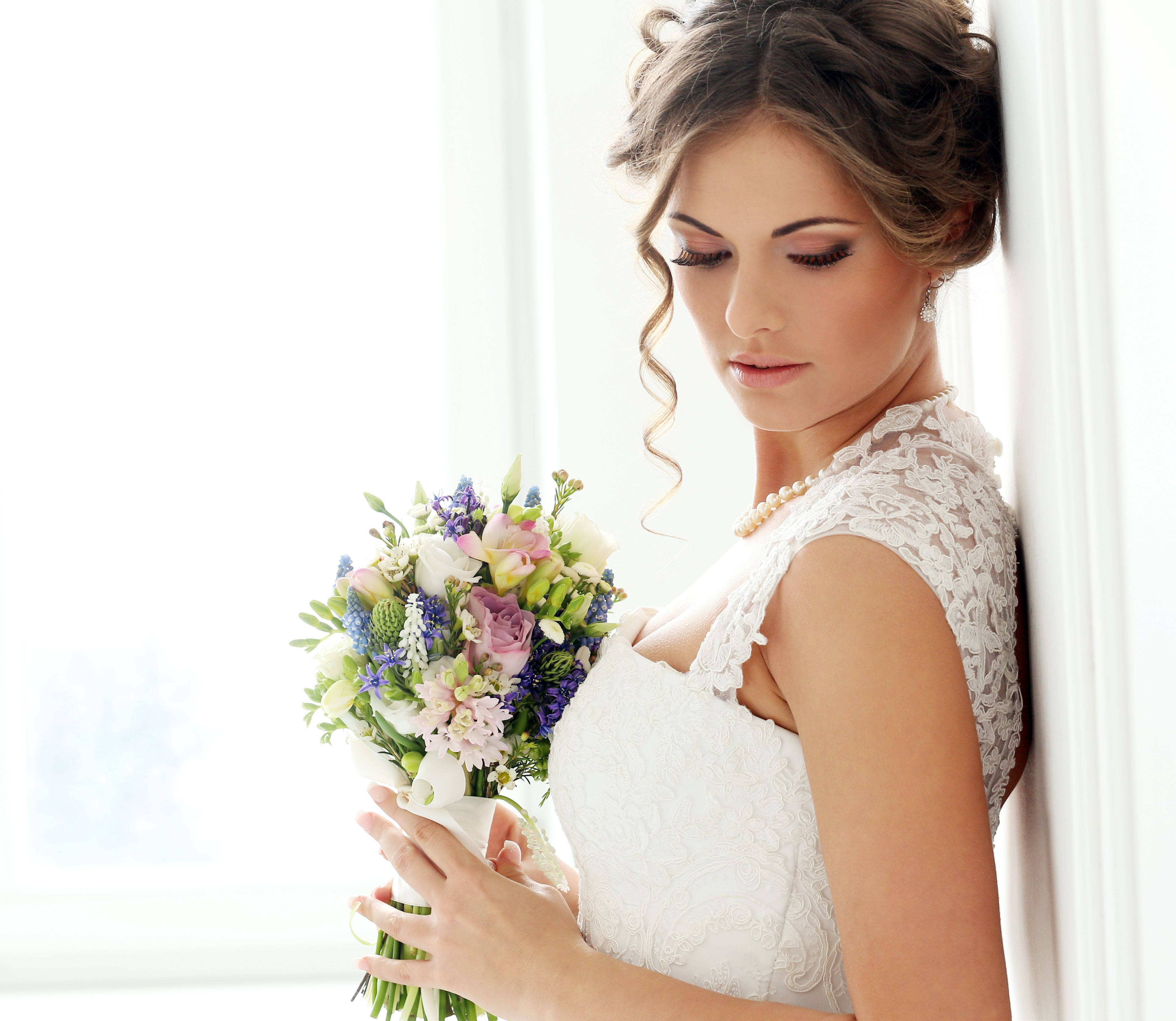 身著精緻婚紗的優雅新娘 壁紙 — 一位身穿白色蕾絲婚紗的新娘靠著明亮的窗戶，手持一束色彩繽紛的花束。