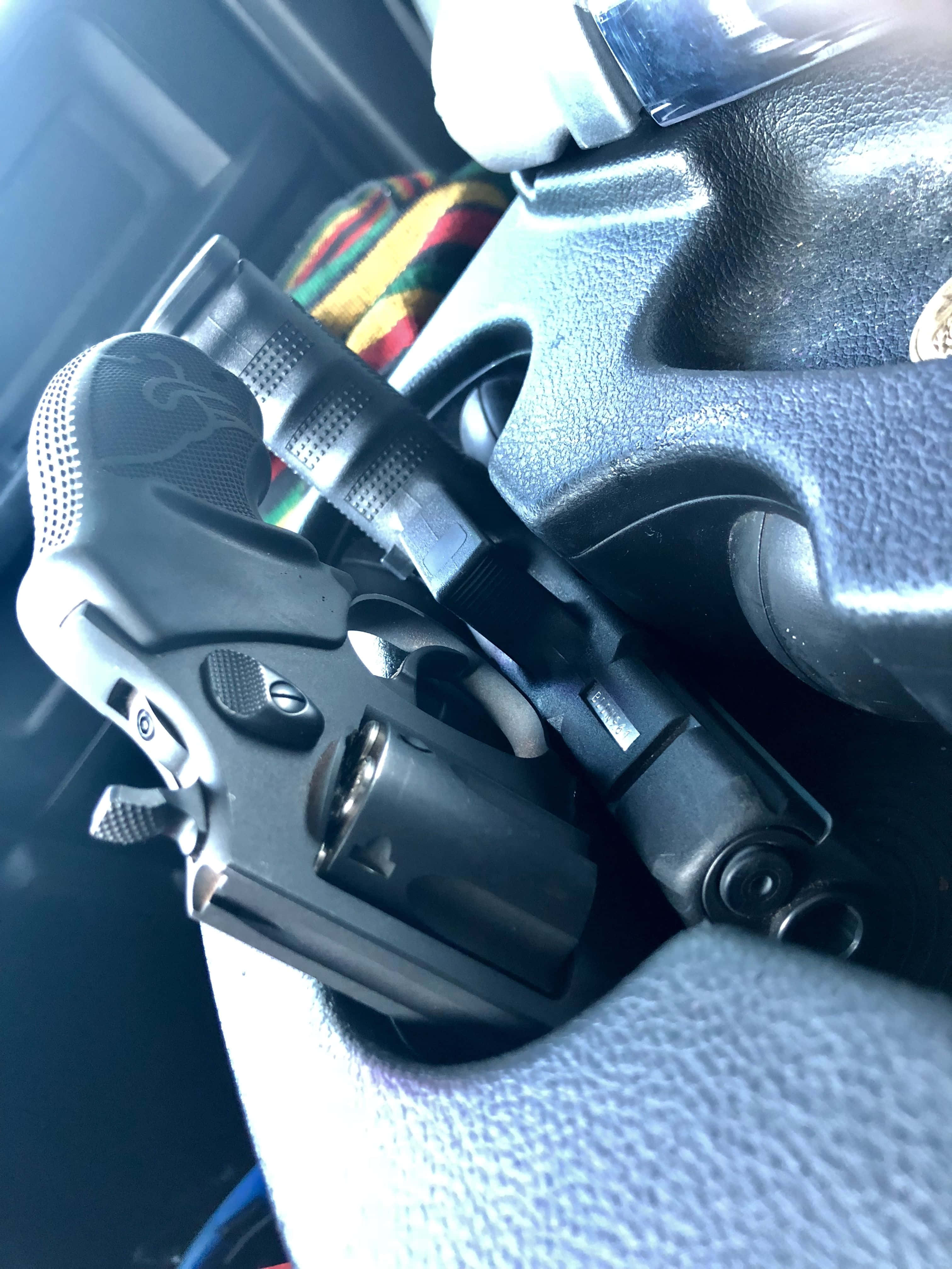 車內手槍圖像 壁紙 — 一把黑色左輪手槍的特寫，放在汽車有紋理的方向盤上，背景有一塊色彩繽紛的布。