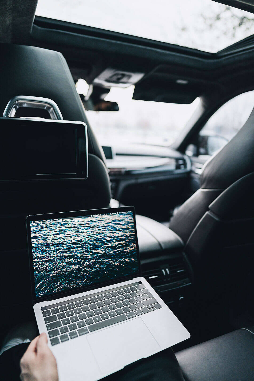 車內的 MacBook Air 2020 壁紙 — 一隻手拿著一部手提電腦，顯示海洋波浪，位於一輛時尚黑色汽車內，天窗顯示模糊的樹木。