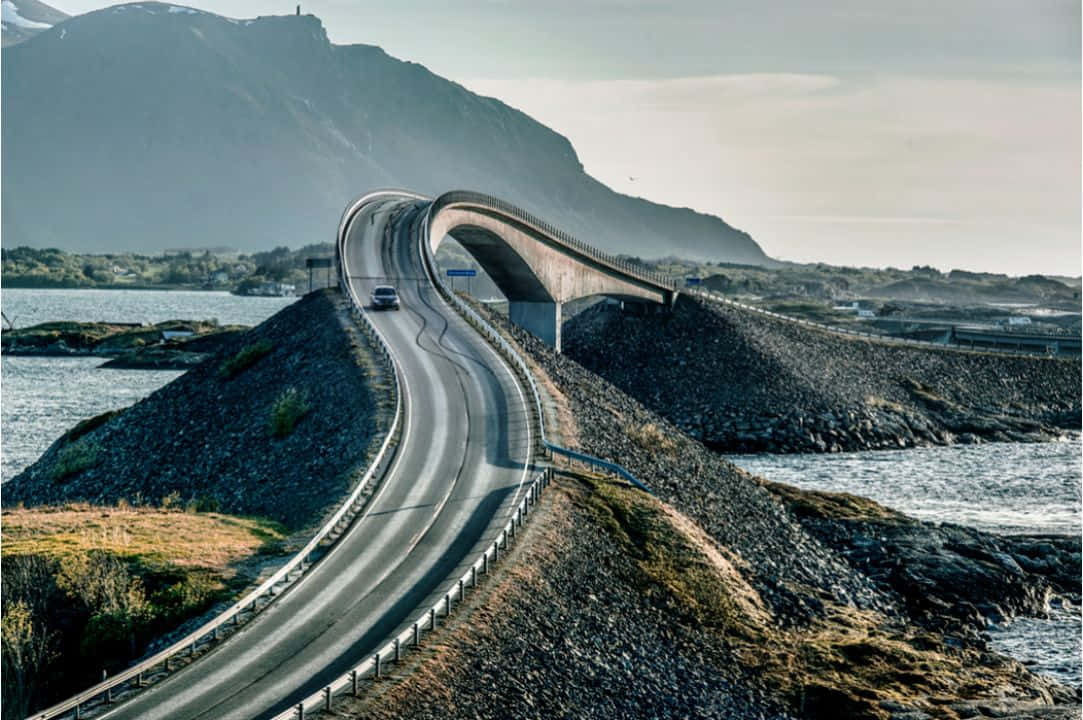 一座彎曲的現代橋樑橫跨水面，一輛汽車在靠近山脈的蜿蜒道路上行駛。