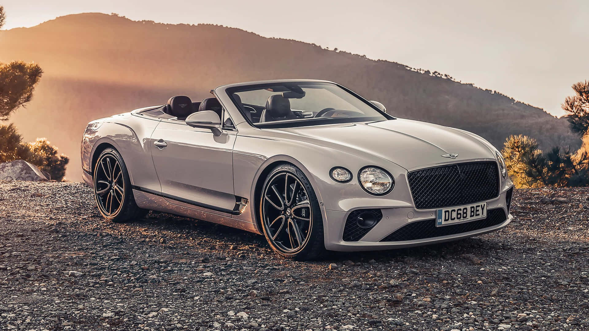 銀色賓利開篷車停泊在碎石上，背景為日落時分的群山。