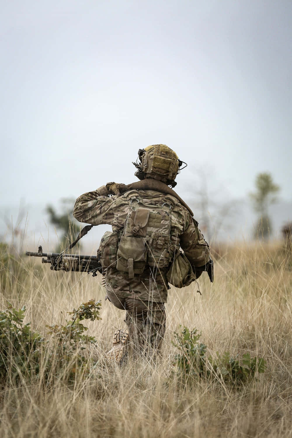 軍事人員野外作戰 壁紙 — 一名身穿全套迷彩裝備的士兵，在陰天之下，手持機槍穿過高高的乾草。