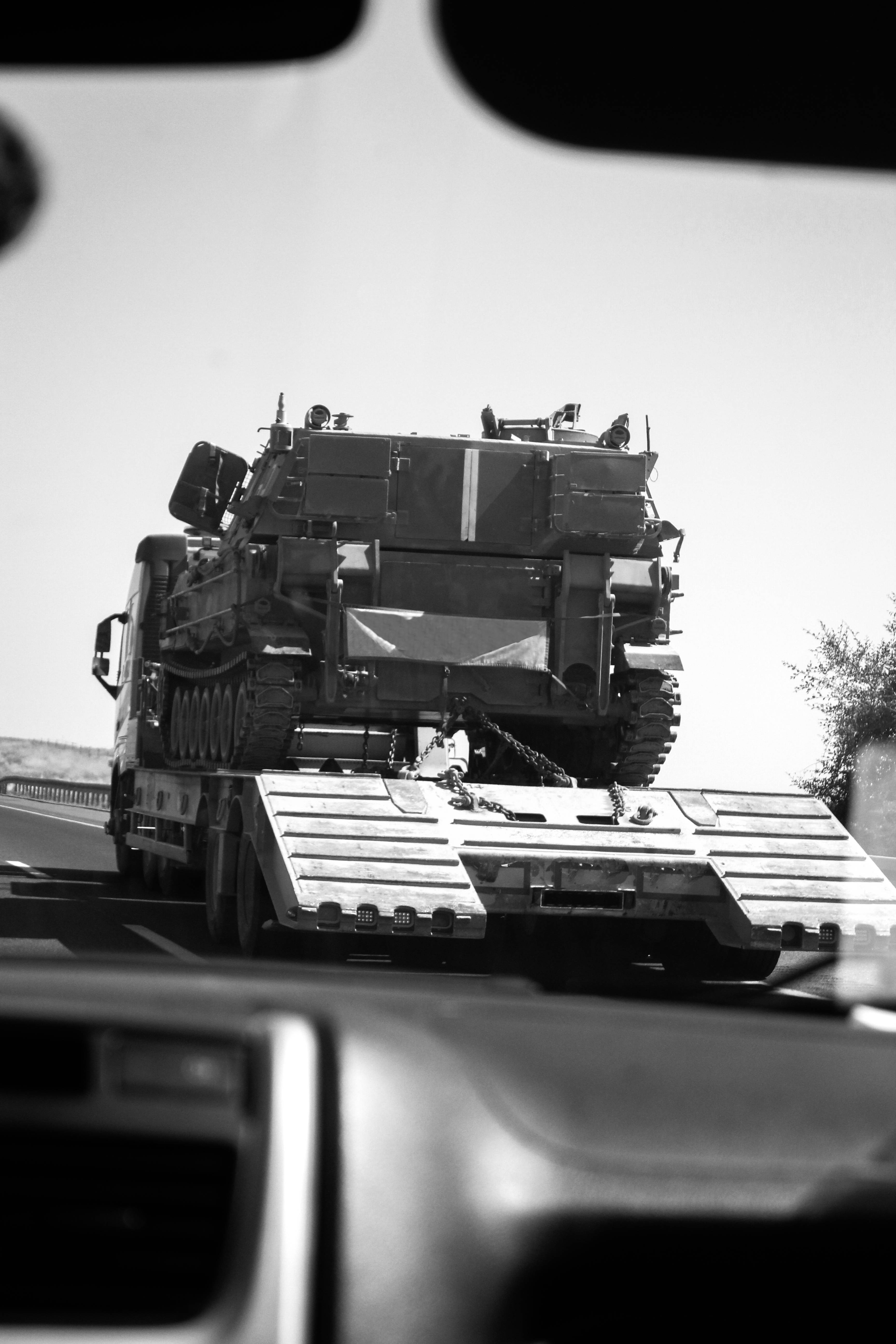 軍用坦克車輛 壁紙 — 從車內拍攝的黑白照片顯示一輛軍用坦克在沙漠高速公路上由平板卡車運輸。