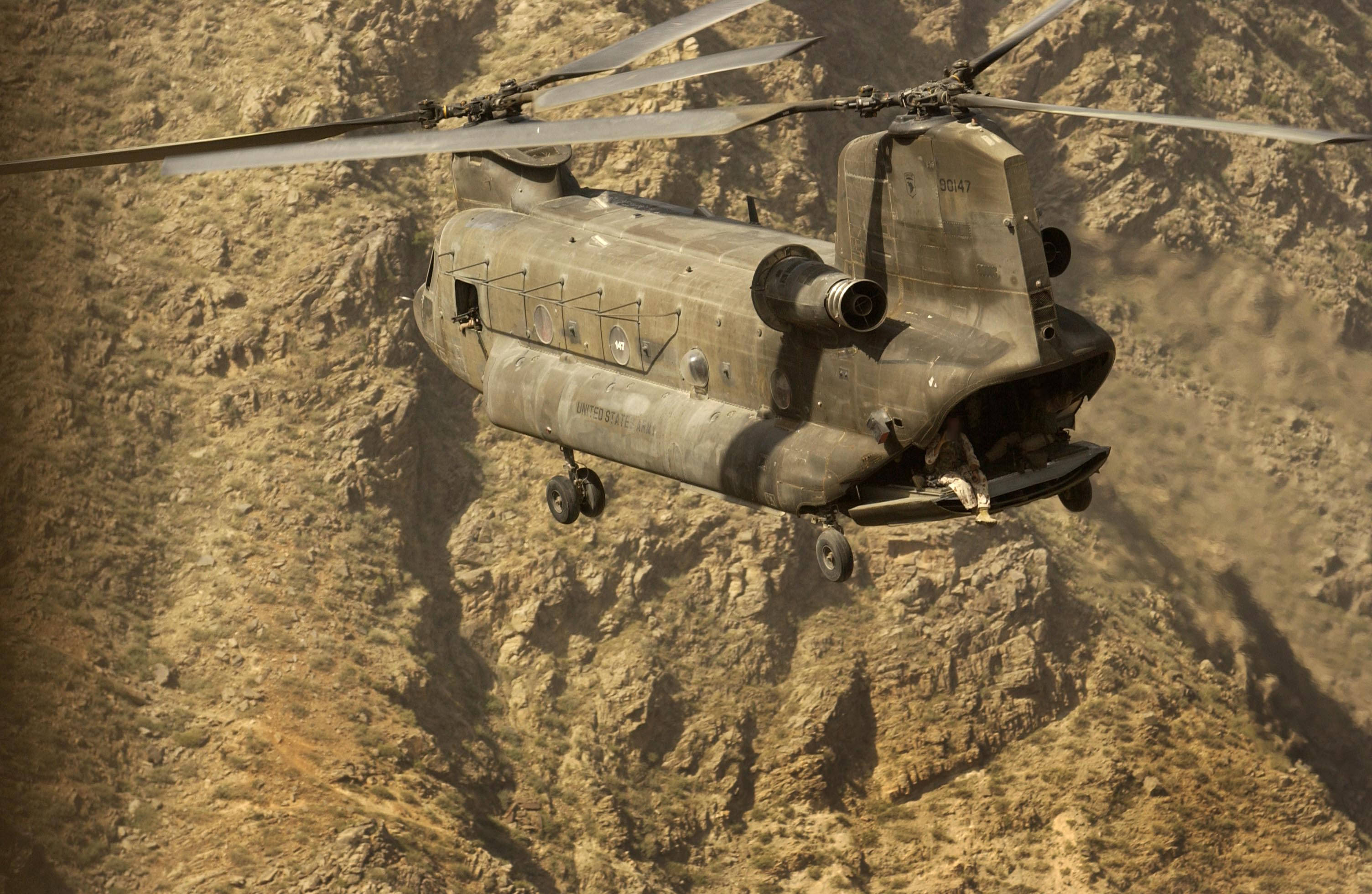 軍用波音Chinook直升機 壁紙 — 一架軍用直升機低飛越過崎嶇的沙漠峽谷地形，揚起下方的灰塵。